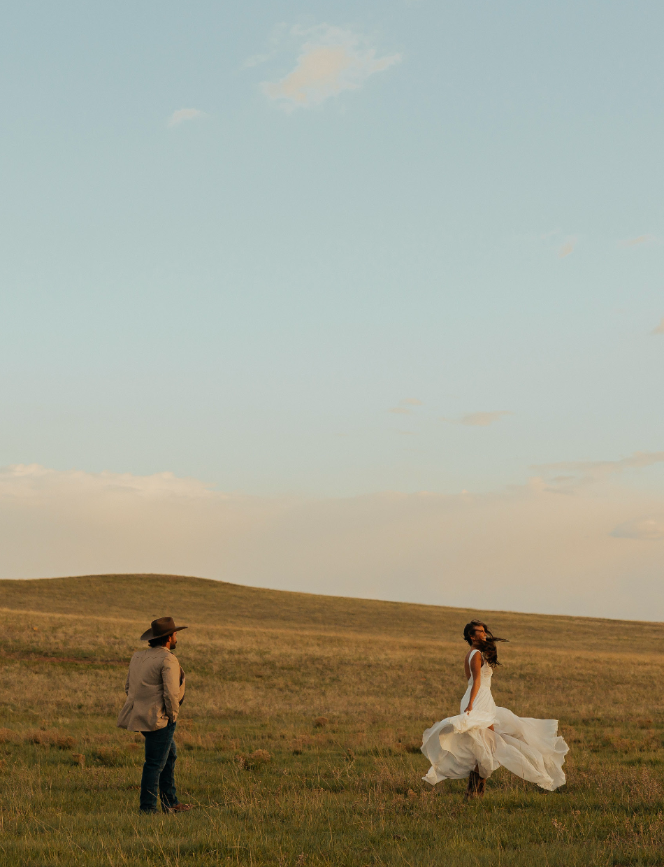 A bride in a flowing white dress and groom in a cowboy hat stand in a vast golden prairie under a soft blue sky.