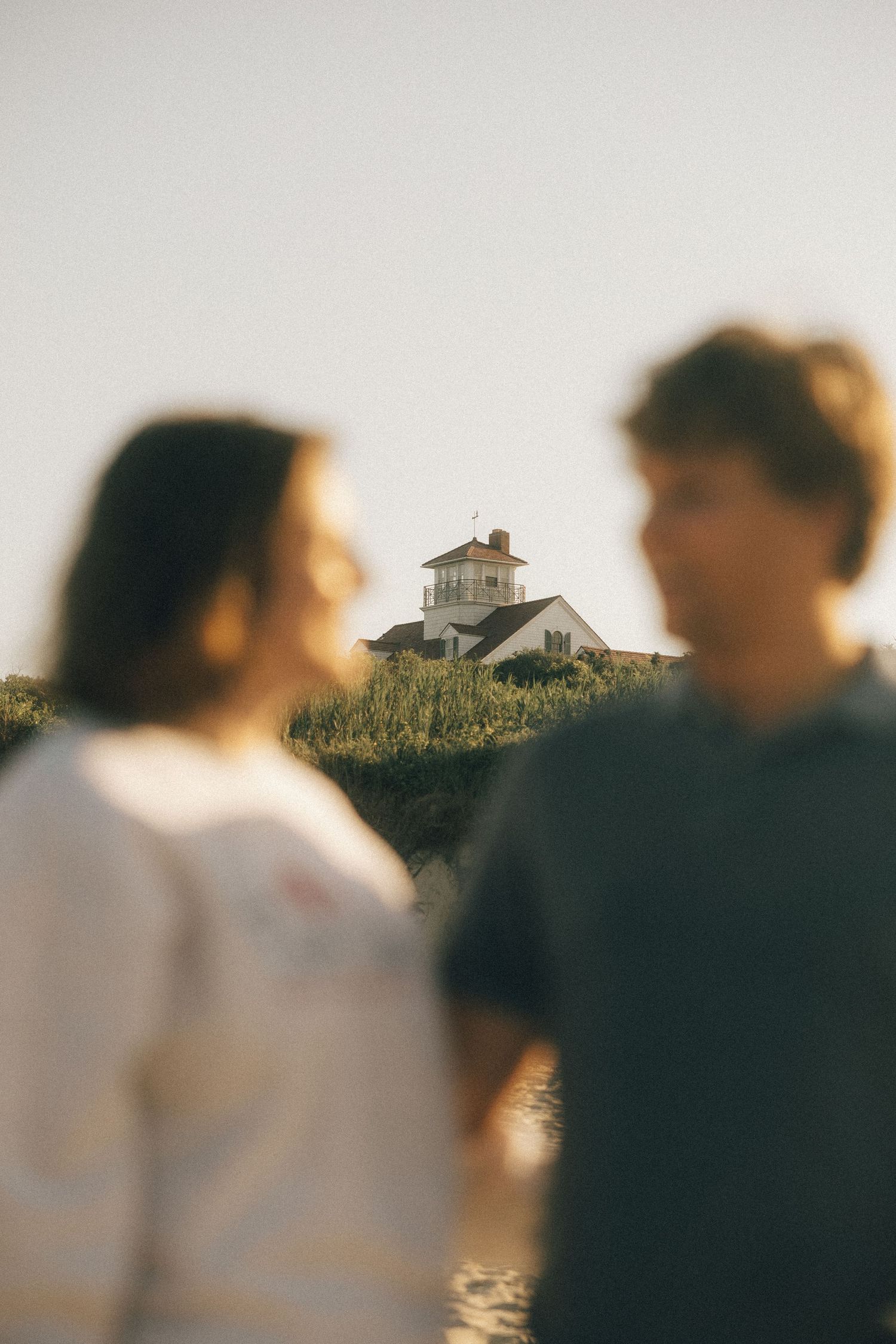 Blurred figures in foreground with coastal house visible in background during sunset.