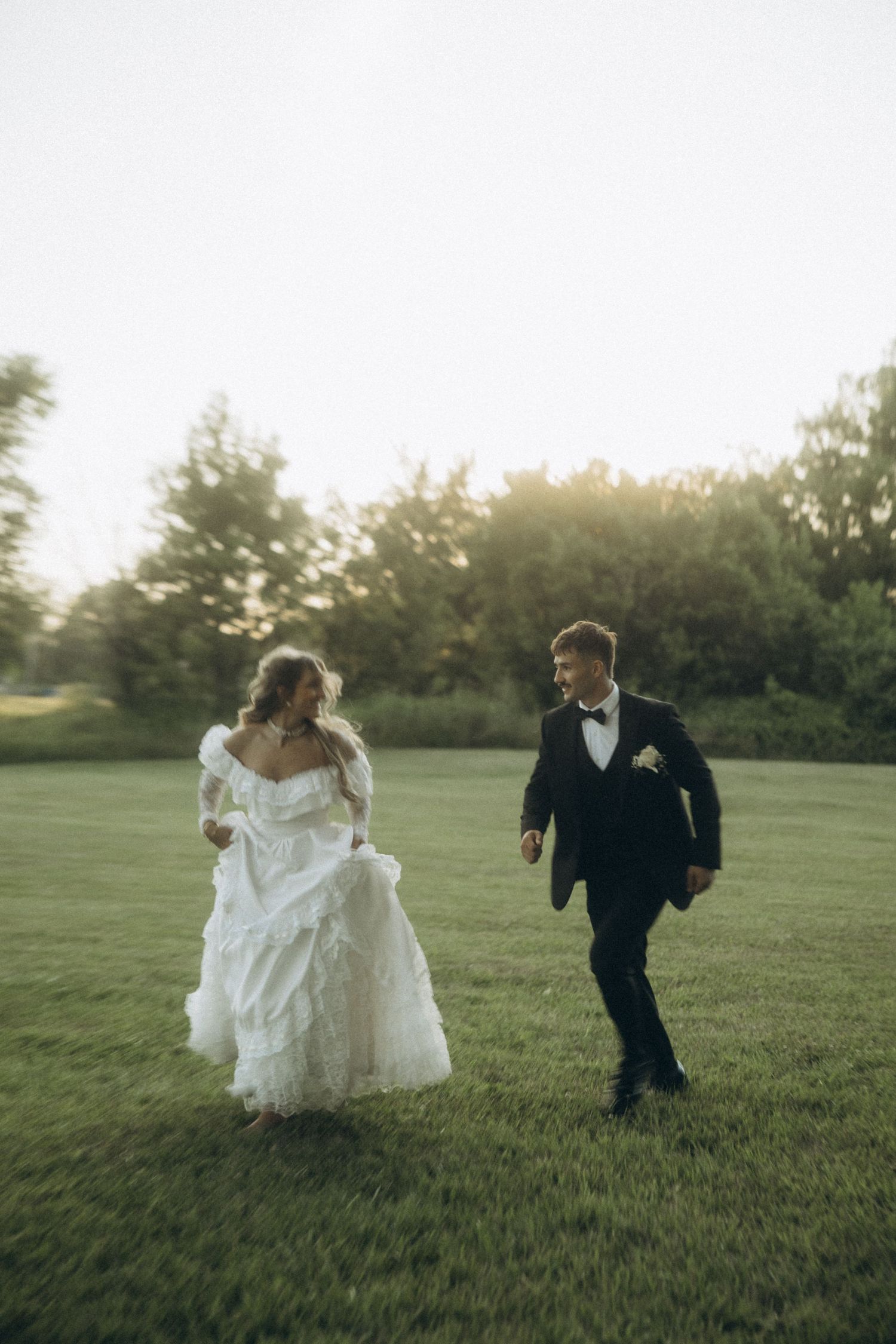 Couple in wedding attire running playfully through a grassy field at sunset.