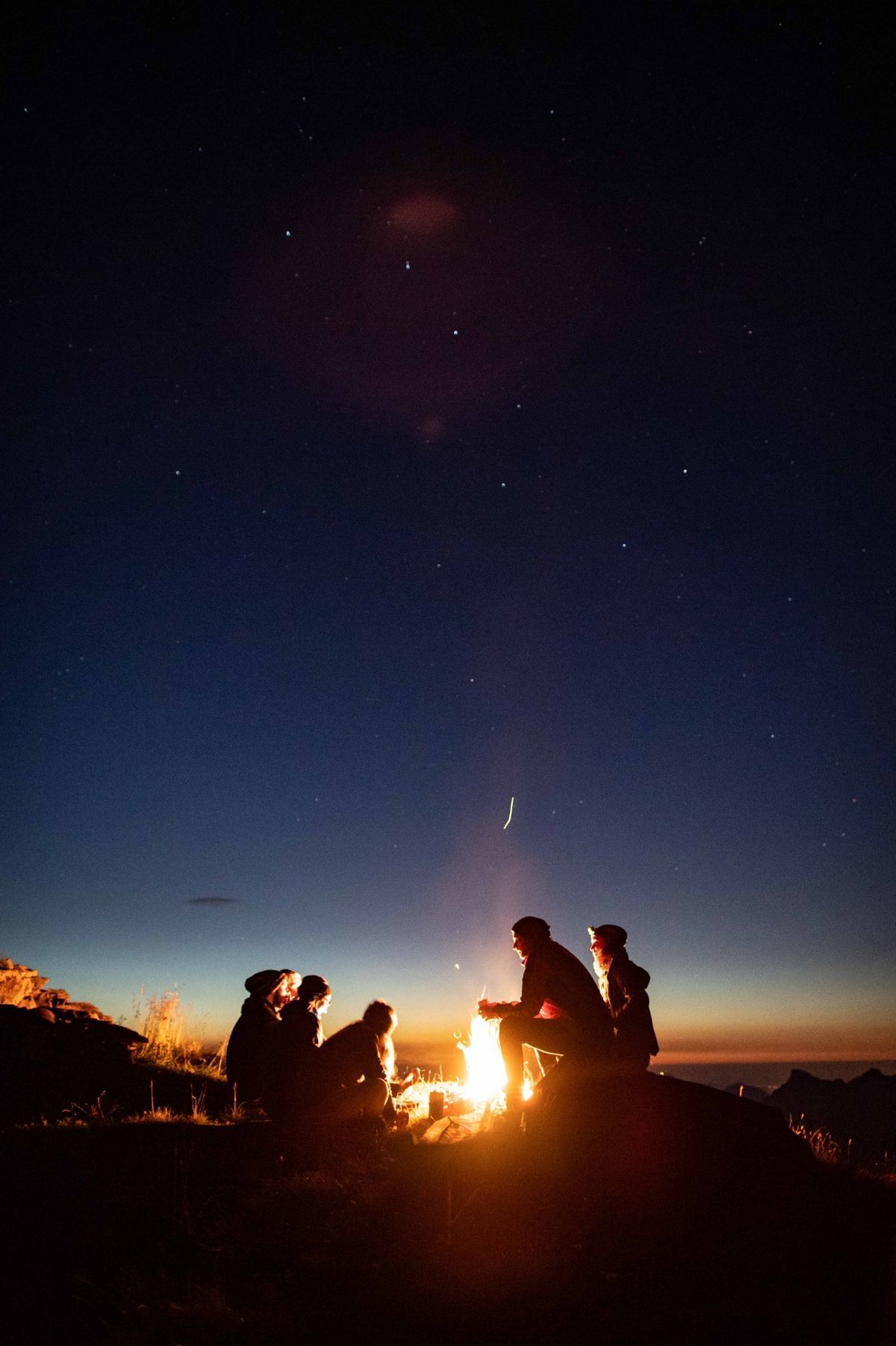 Silhouettes of friends gathered around a campfire under a starry night sky with a glowing horizon.