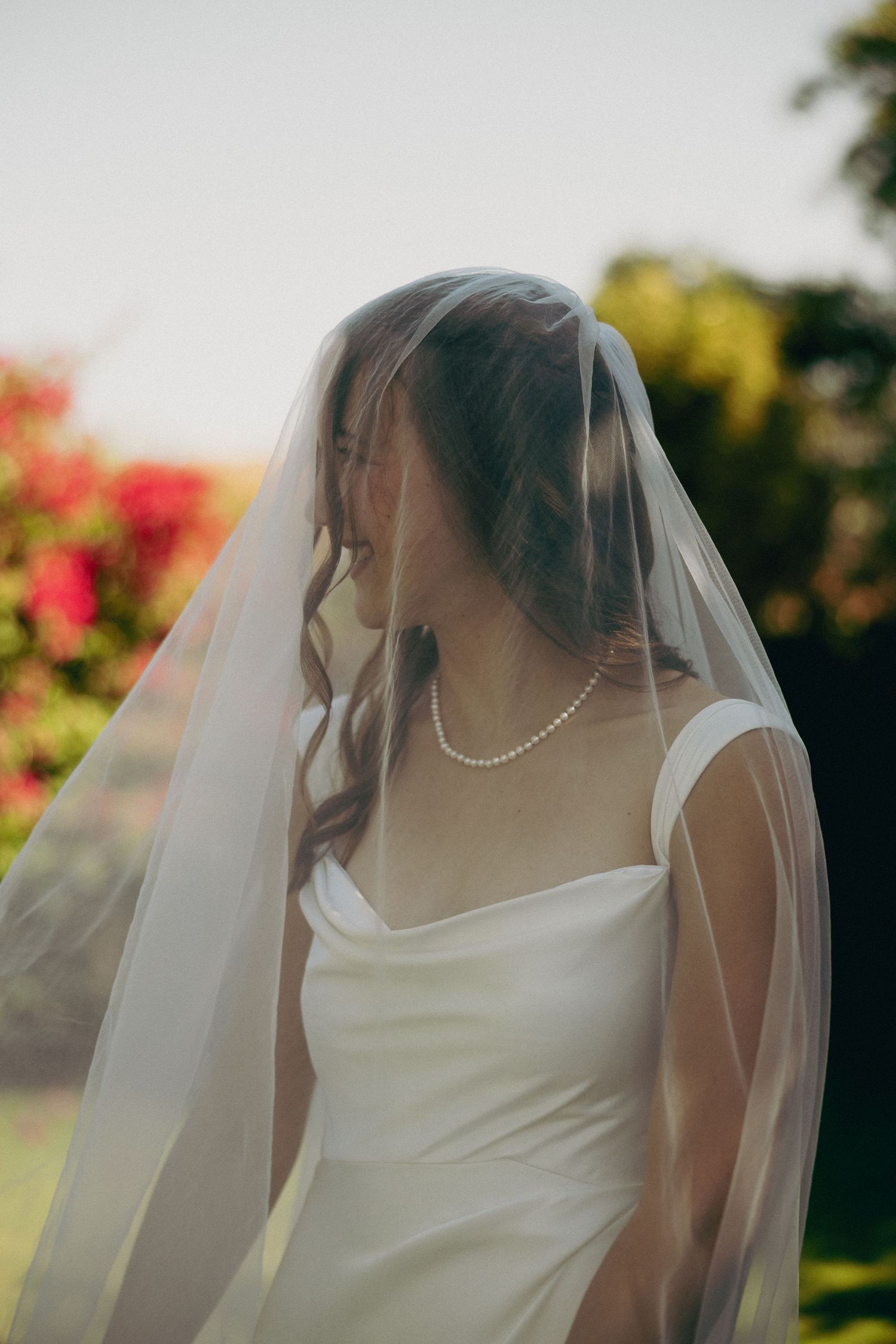 Elegant bride in white dress and veil standing outdoors with blooming flowers in background.