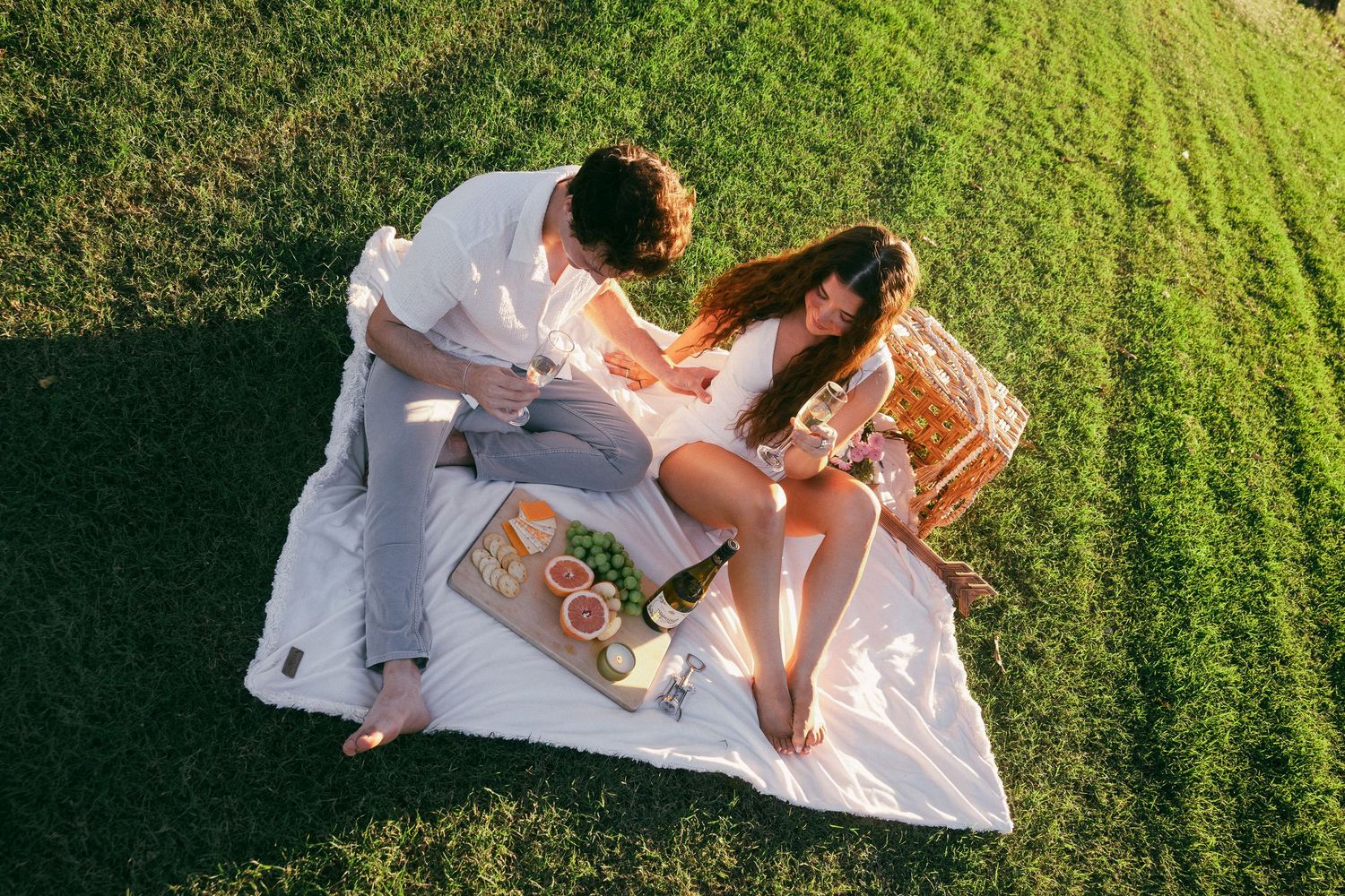 Couple enjoys romantic picnic on white blanket spread across green grass.