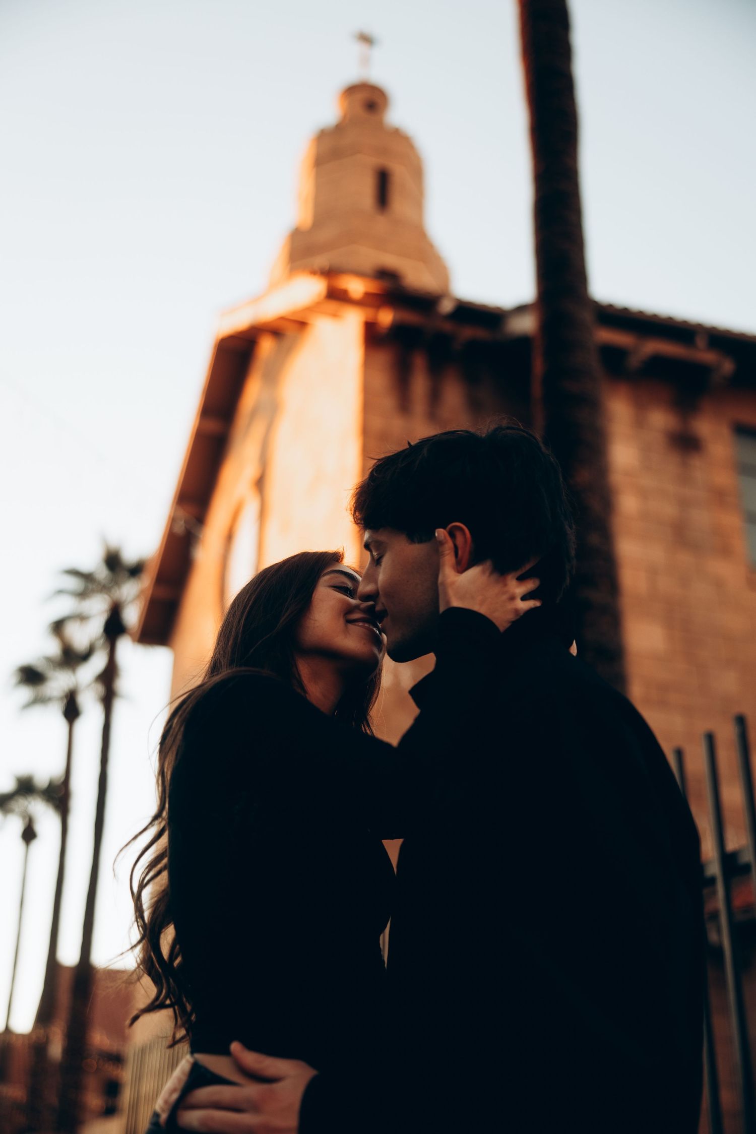 Silhouetted couple embraces at sunset with palm trees and architectural towers in warm golden light.