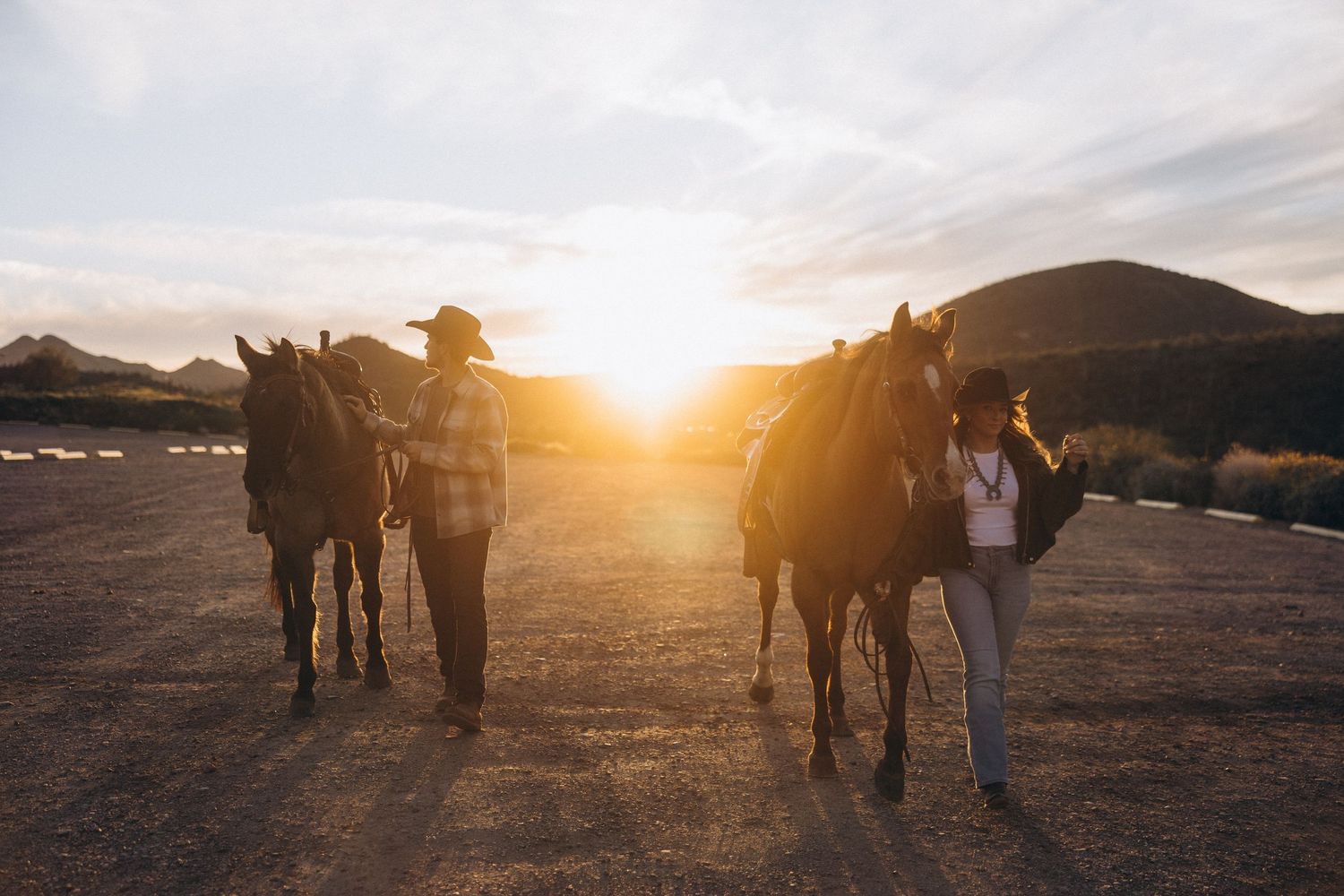 Two people stand with their horses in a desert landscape during golden sunset hour.