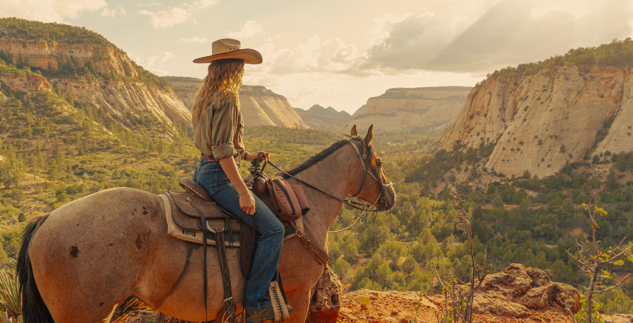 A cowgirl in a hat rides a brown horse overlooking a stunning canyon landscape at golden hour.