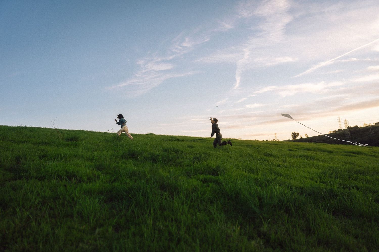 Two people flying a kite on a lush green hillside at sunset under a blue sky.