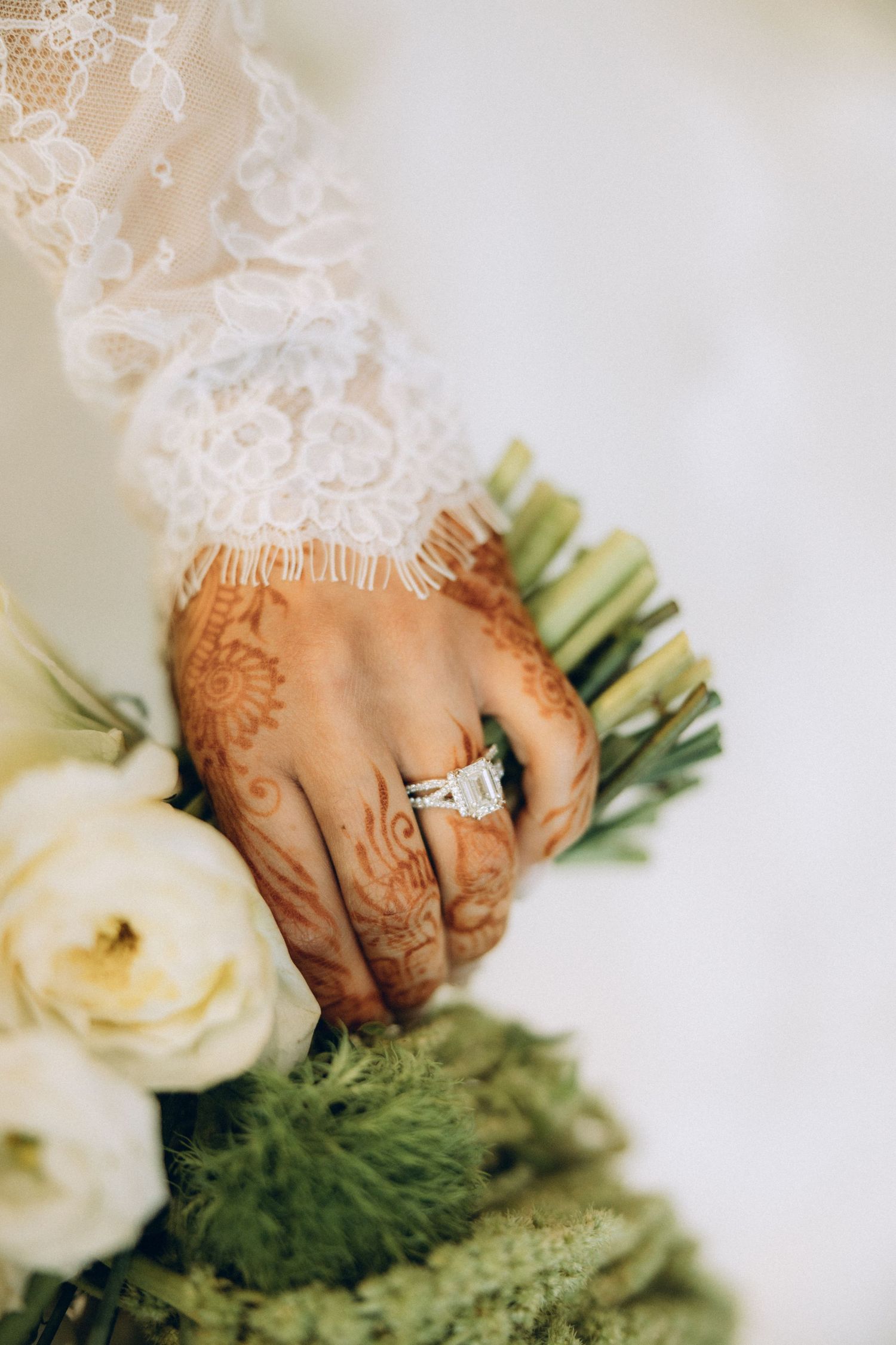Close-up of hands adorned with henna designs holding a bouquet against white lace fabric.