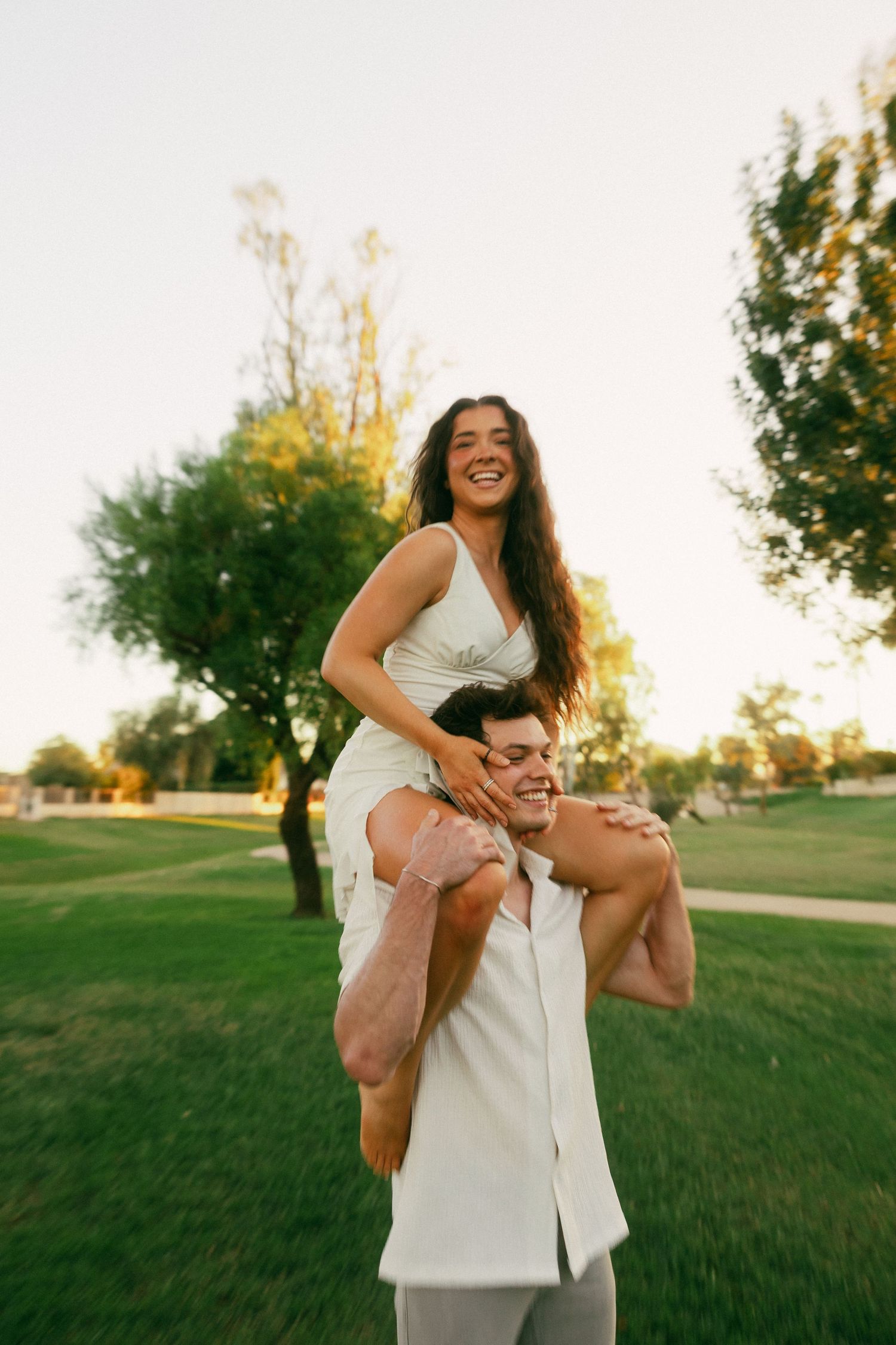 Happy couple in a sunny park, woman sitting on man's shoulders, both smiling, wearing white outfits.