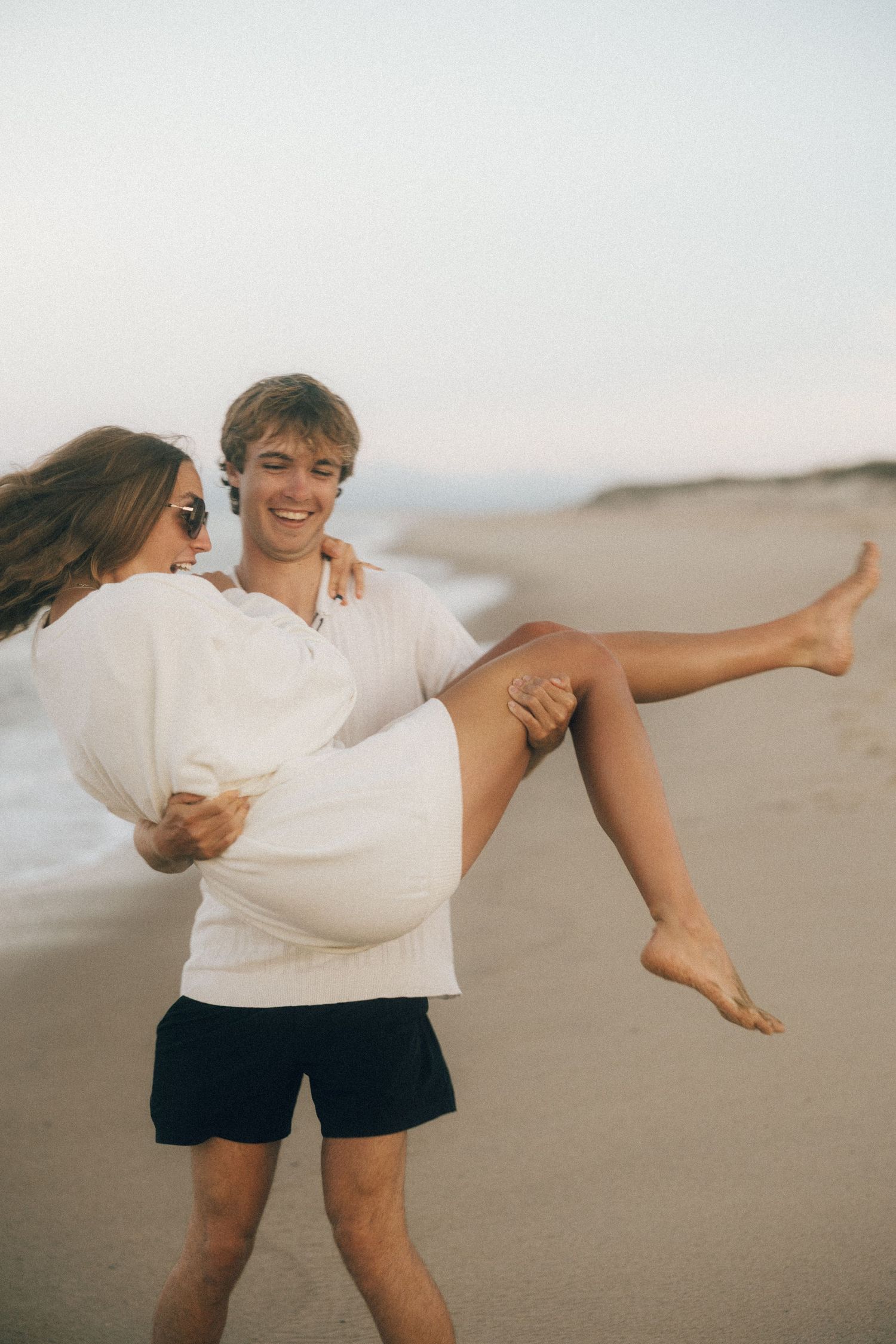 A smiling man carries a laughing woman in his arms on a sandy beach at dusk, both wearing white tops.