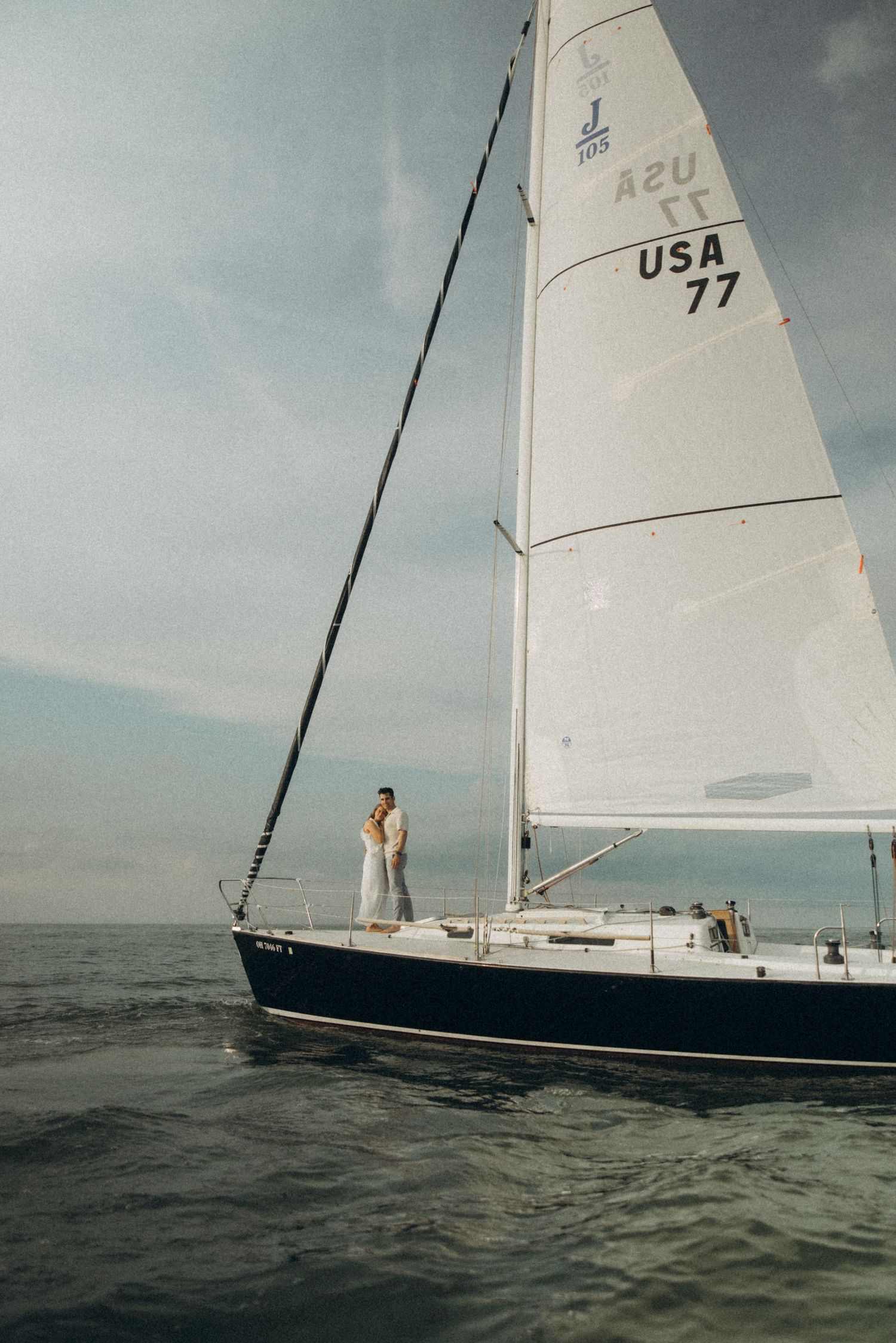 A sleek sailboat with white sails glides across dark waters under cloudy skies.