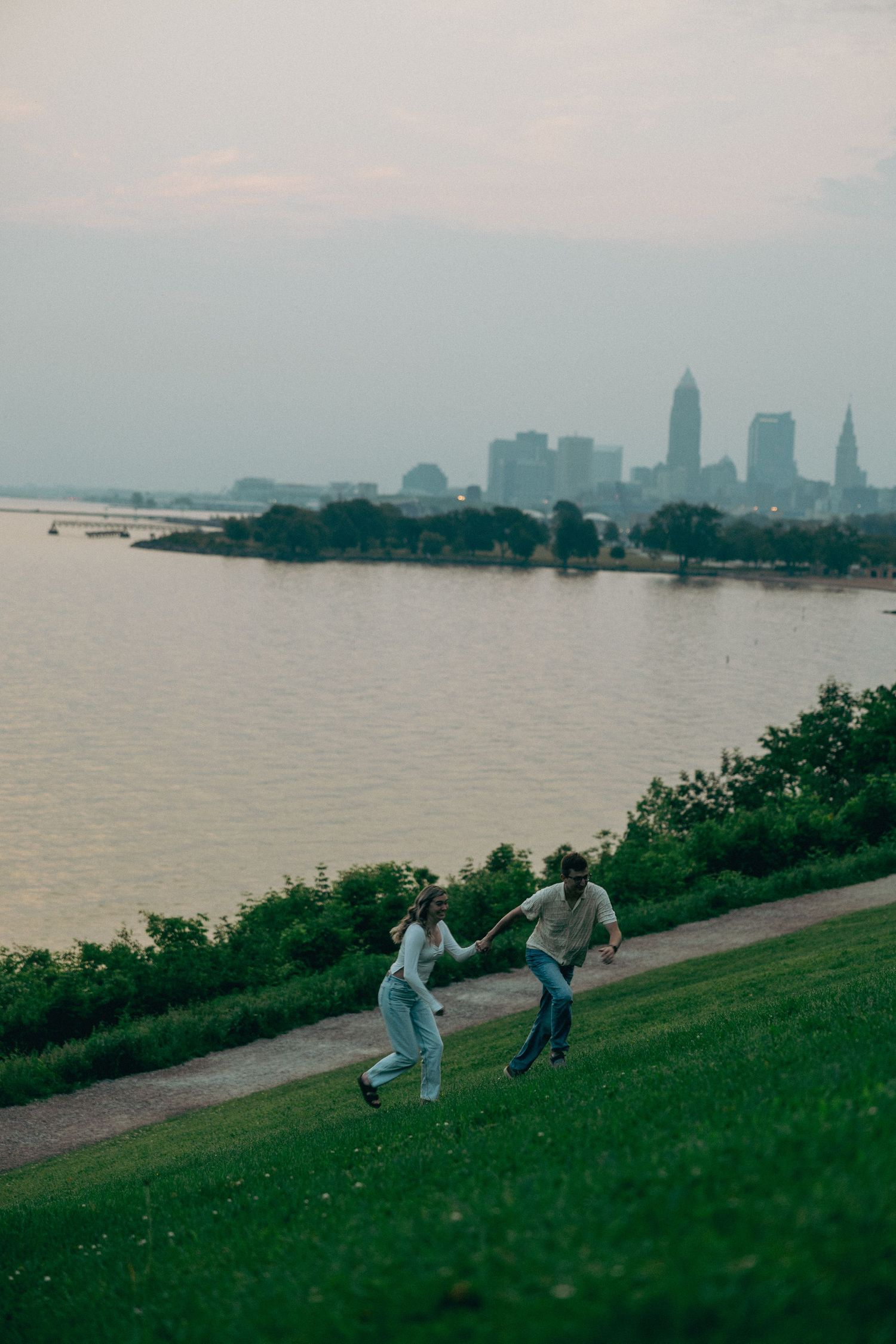 Couple playfully running together on a grassy hill overlooking a city skyline and waterfront.