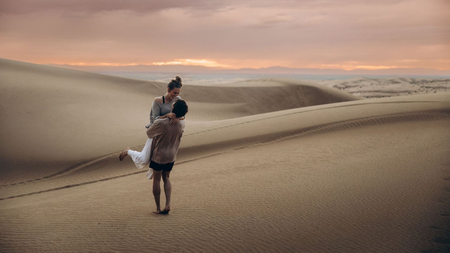 A couple embraces in a romantic moment on rolling sand dunes at sunset with soft pink and orange sky.