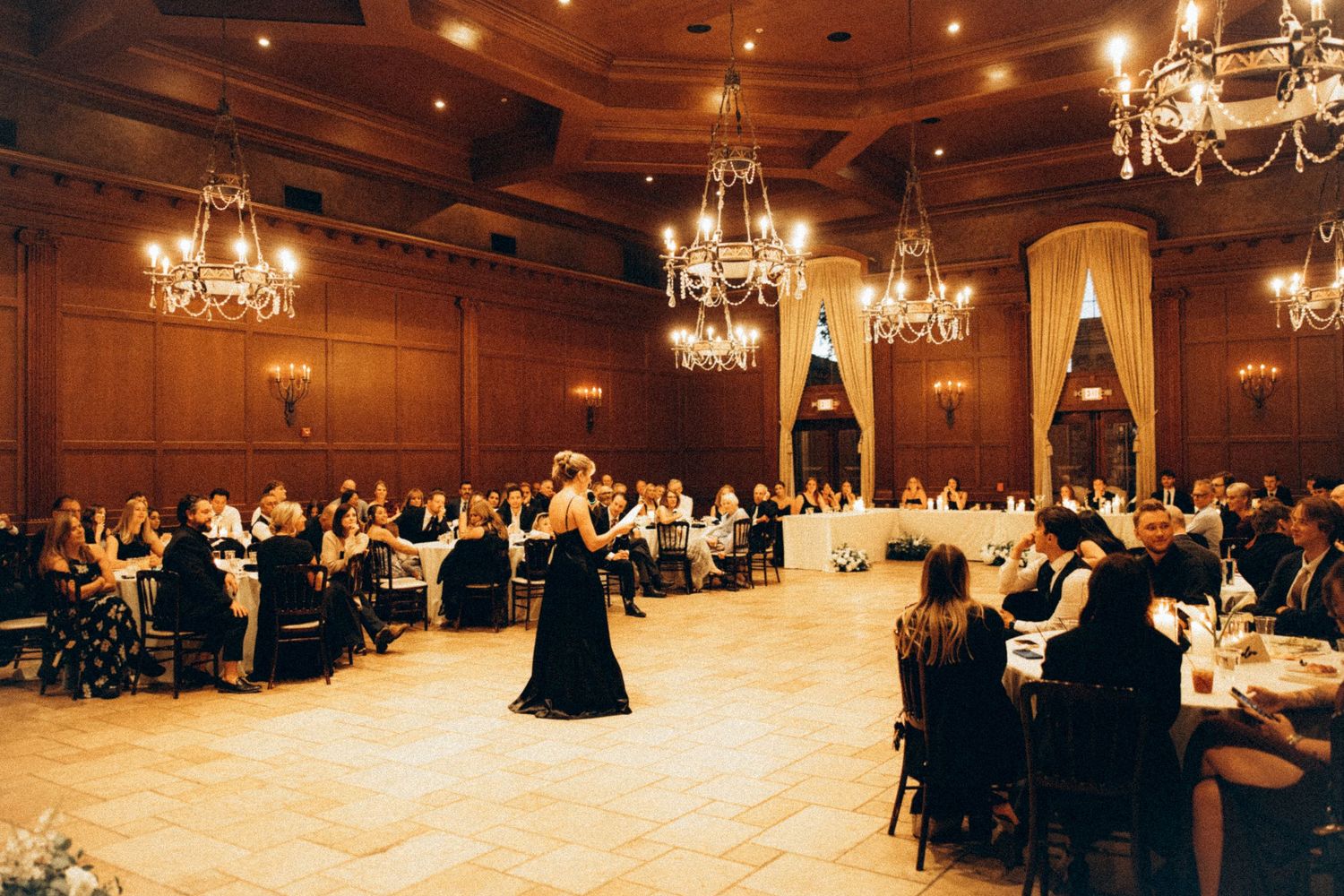 Elegant ballroom wedding reception with chandeliers, wood-paneled walls, and guests seated at round tables while a performer entertains.