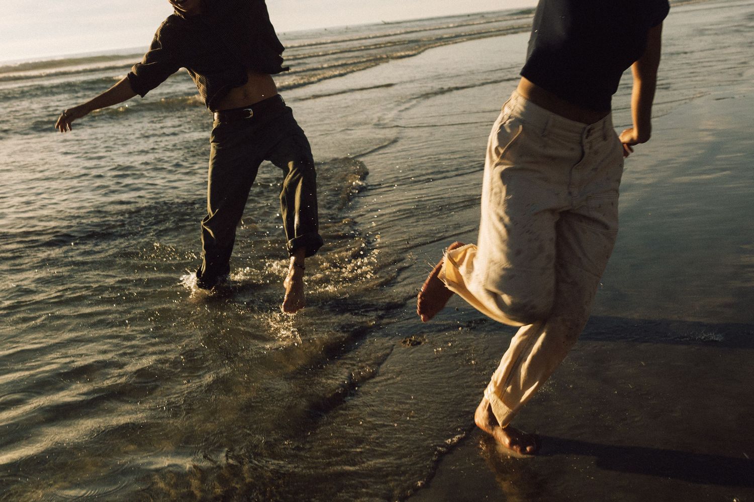 Two people walking barefoot in shallow water on a sandy beach at sunset, creating ripples in the calm water.
