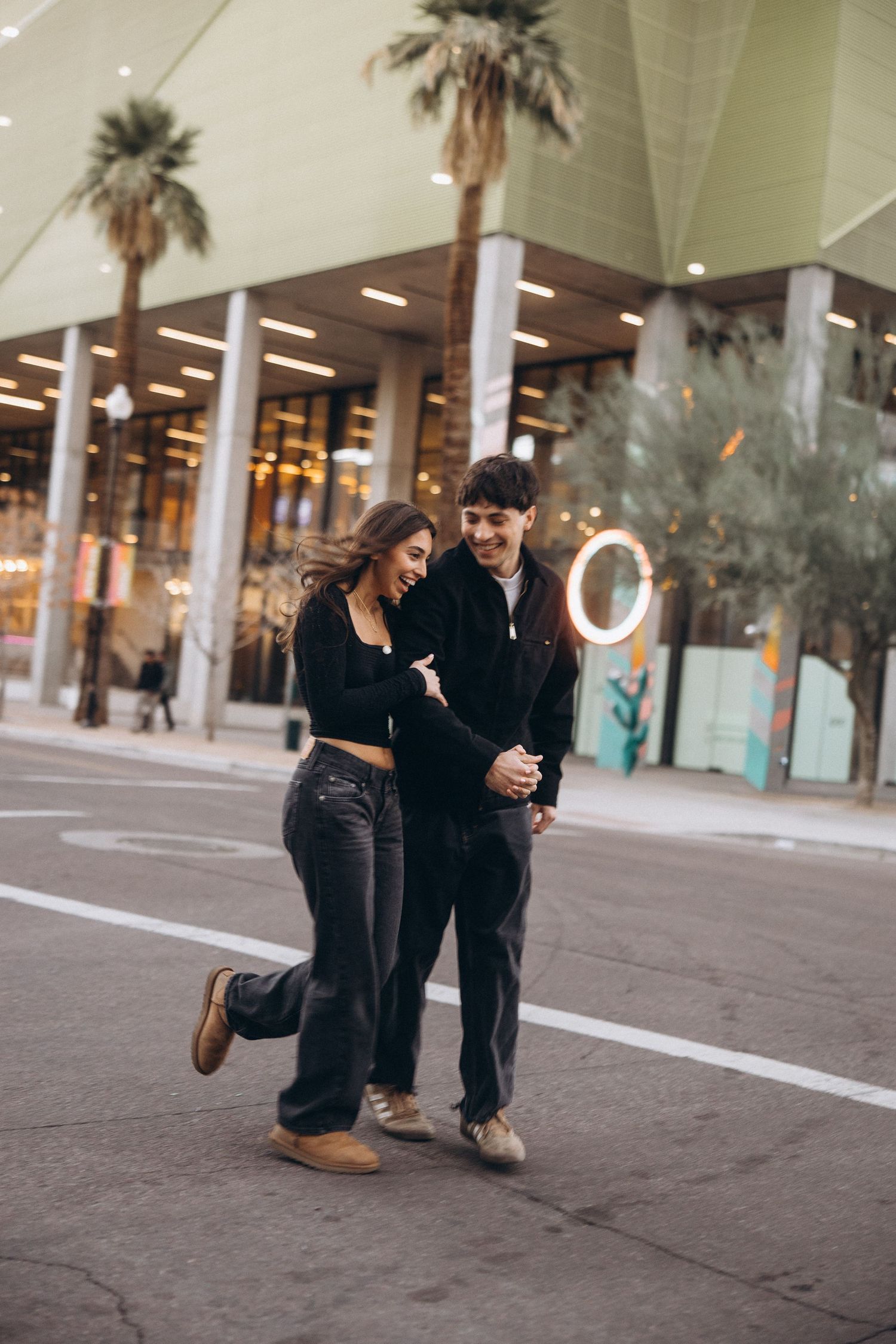 A couple in dark clothing shares a tender moment on a palm-lined street near storefronts at dusk.