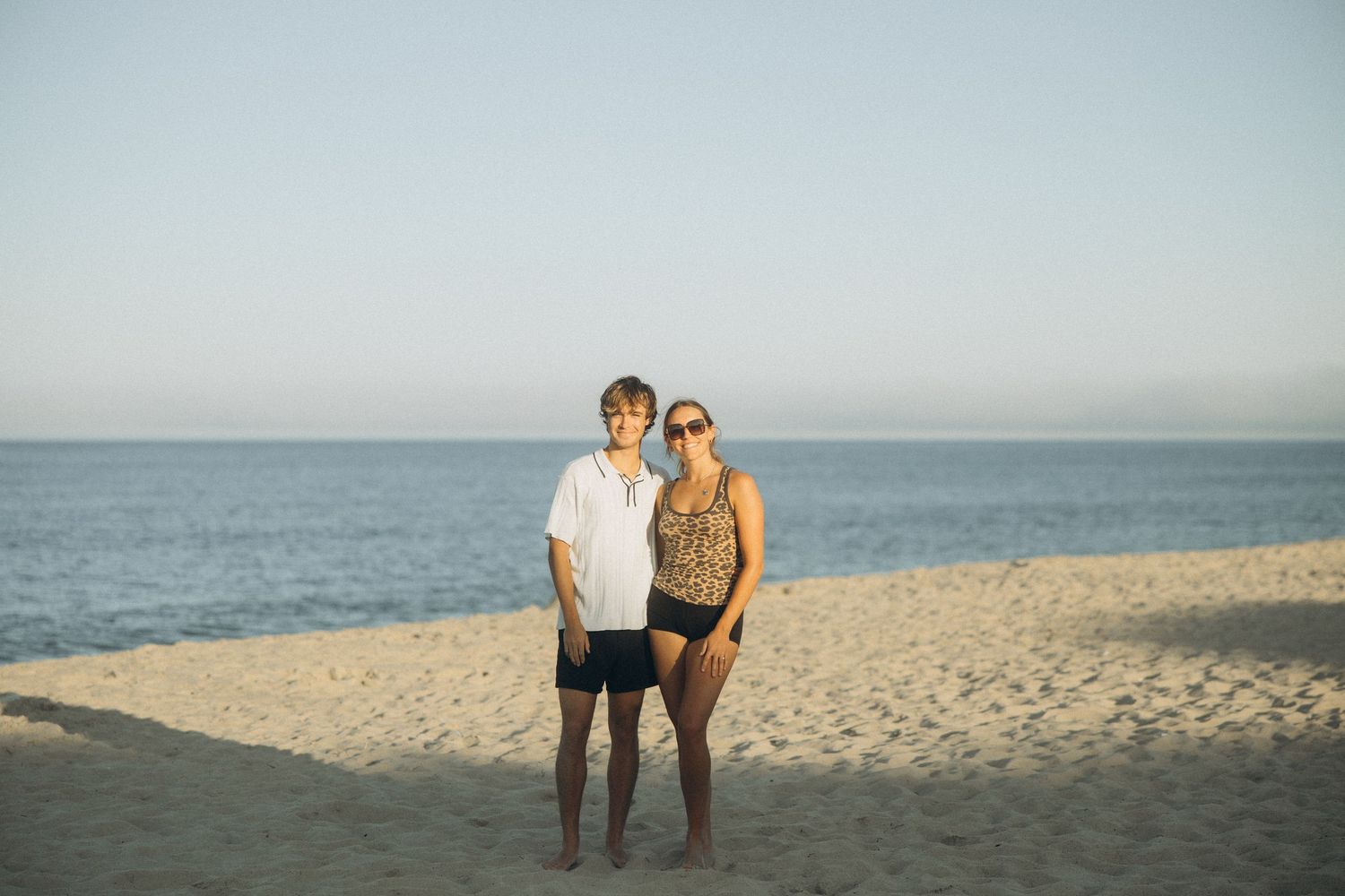 Couple standing together on a sandy beach with calm ocean and clear sky in the background.