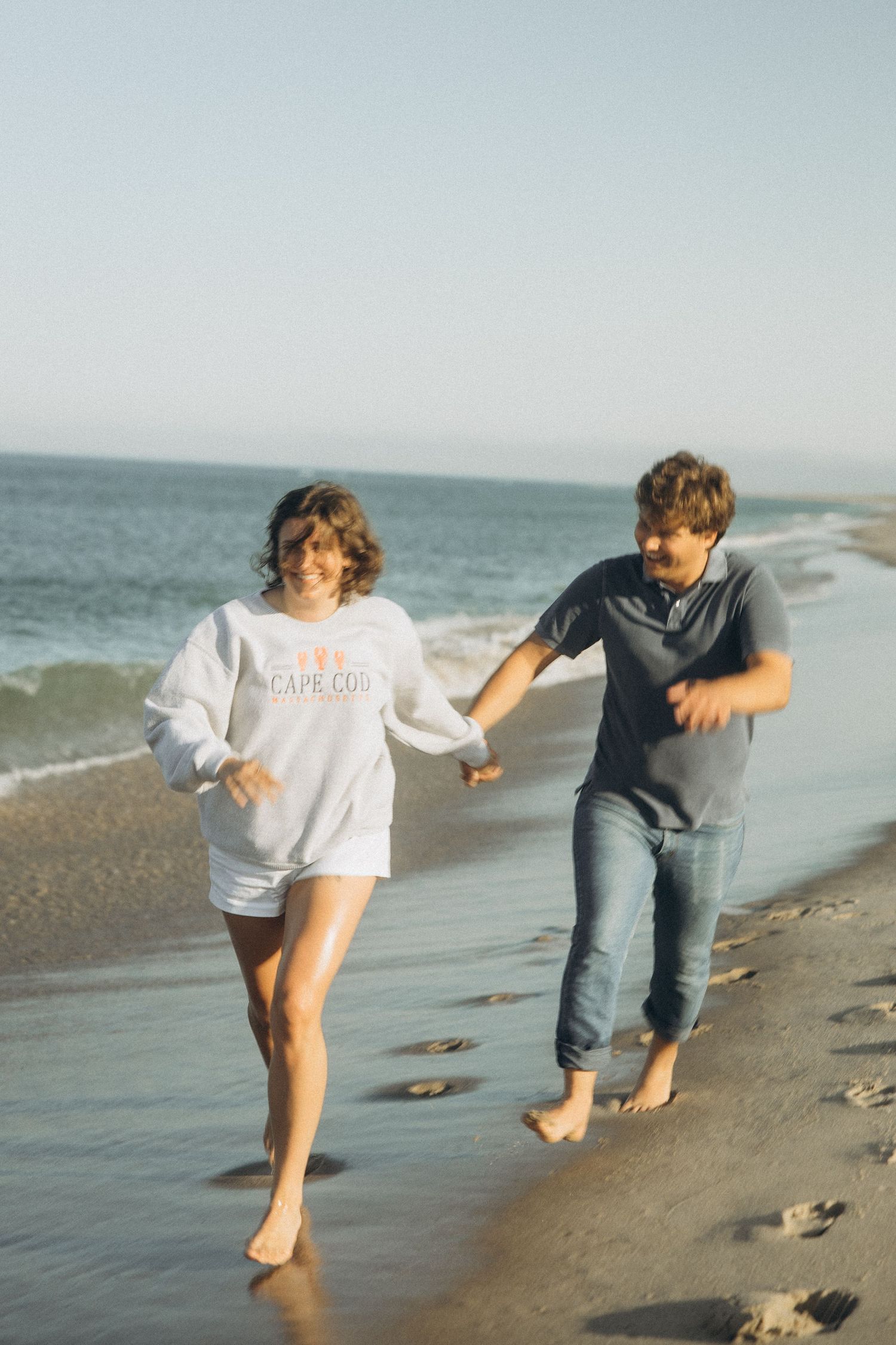 Two people playfully run and jump along a sandy beach by the ocean.