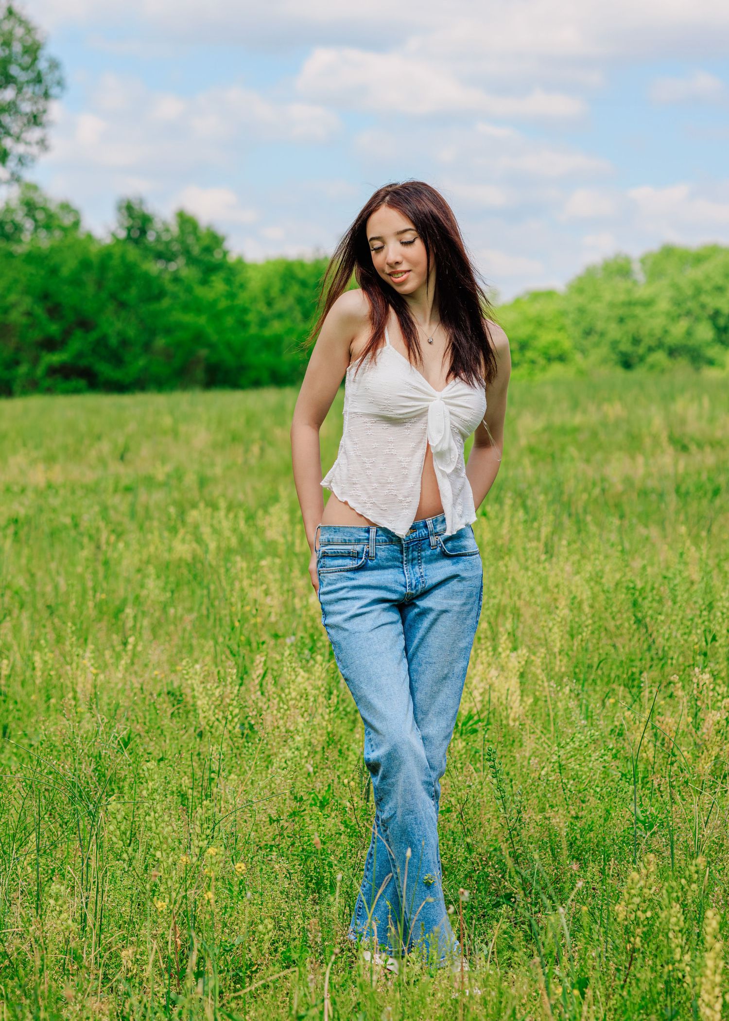 Young woman in white top and blue jeans standing in a lush green meadow on a sunny day.