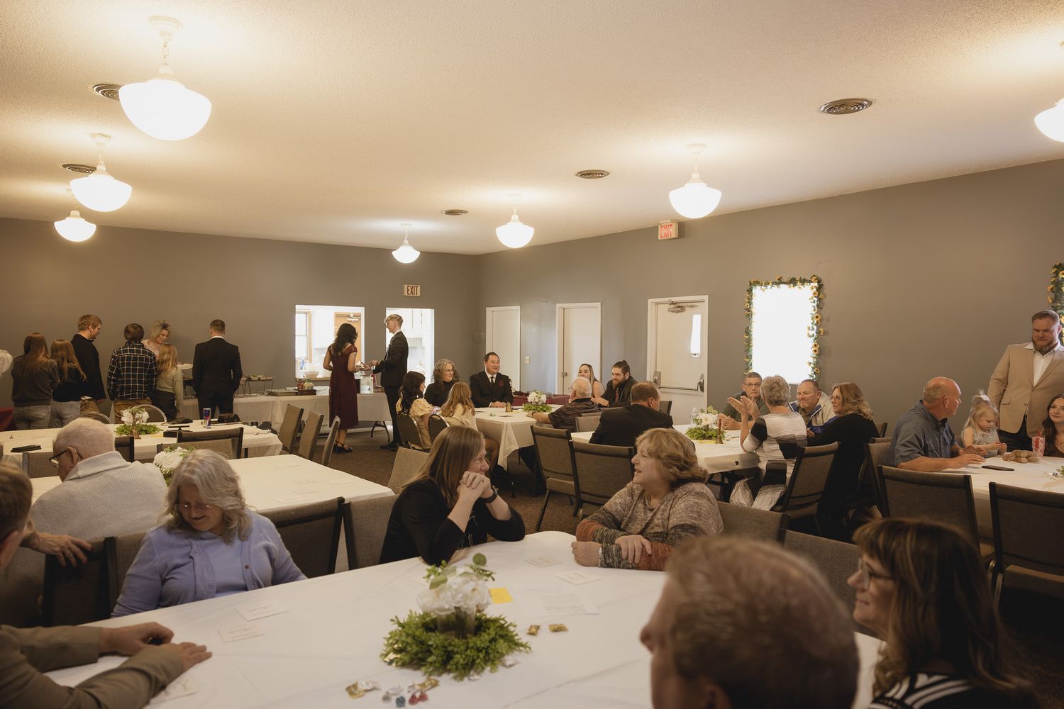 A gathering in a reception hall with round tables and ceiling lights where people are mingling.