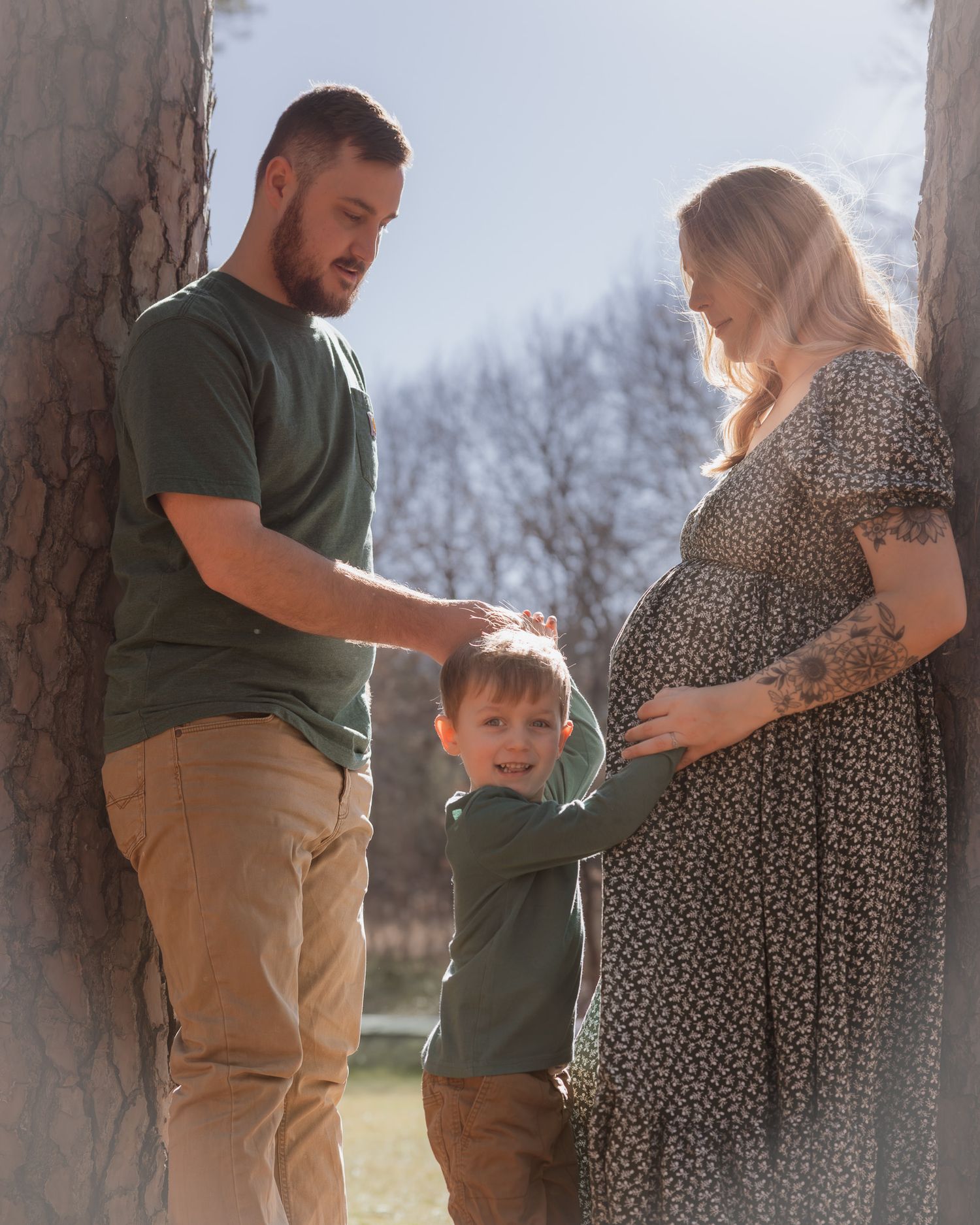 A couple and child pose together against tree trunks in a natural outdoor setting during golden hour.