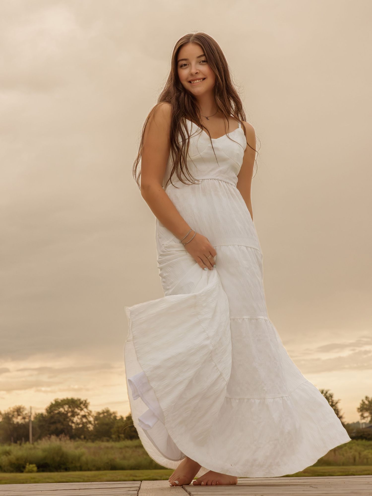 Young woman in a flowing white maxi dress standing outdoors at sunset with a golden sky backdrop.