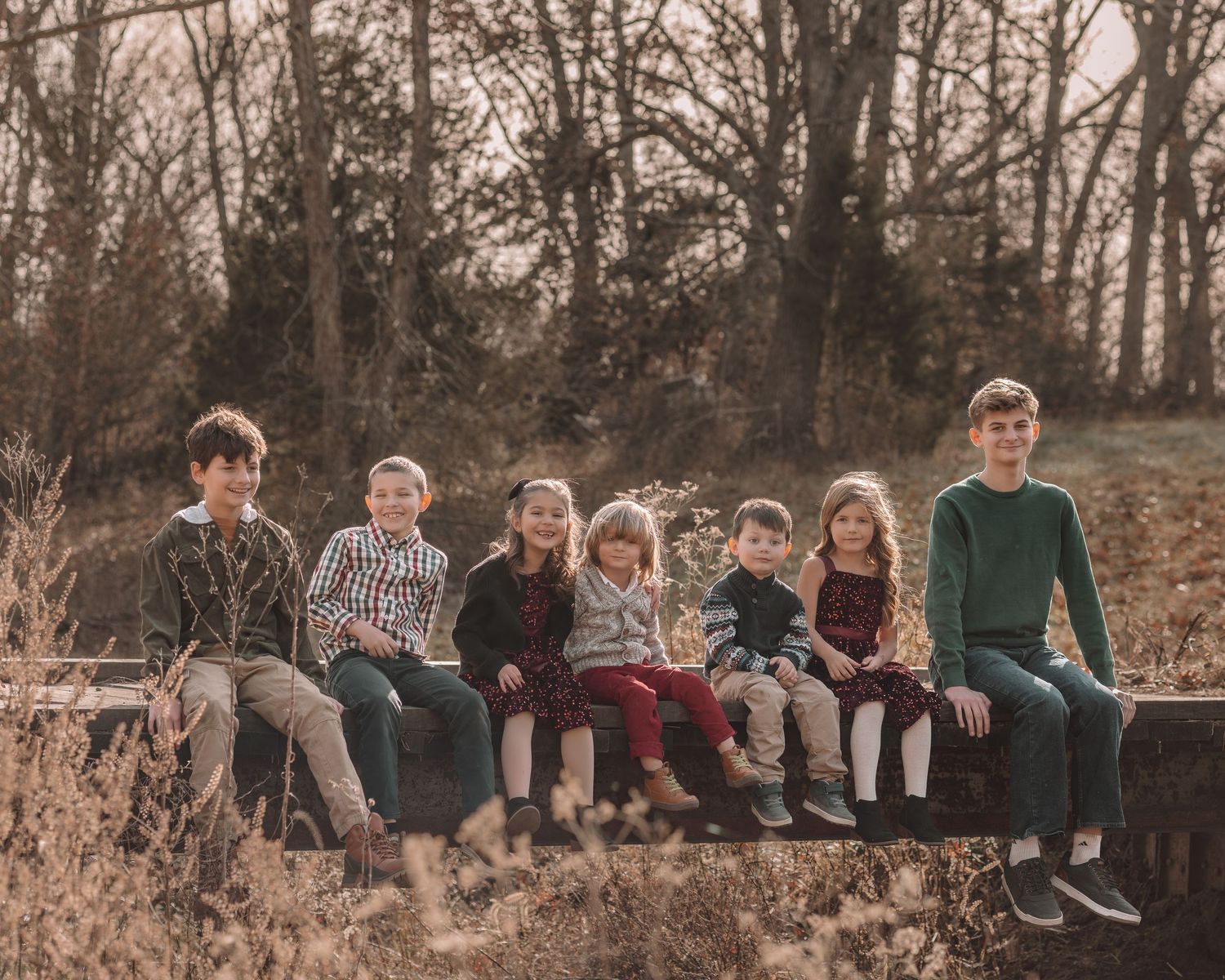 Group of people sitting casually on a rustic wooden fence in an autumn forest setting at sunset.