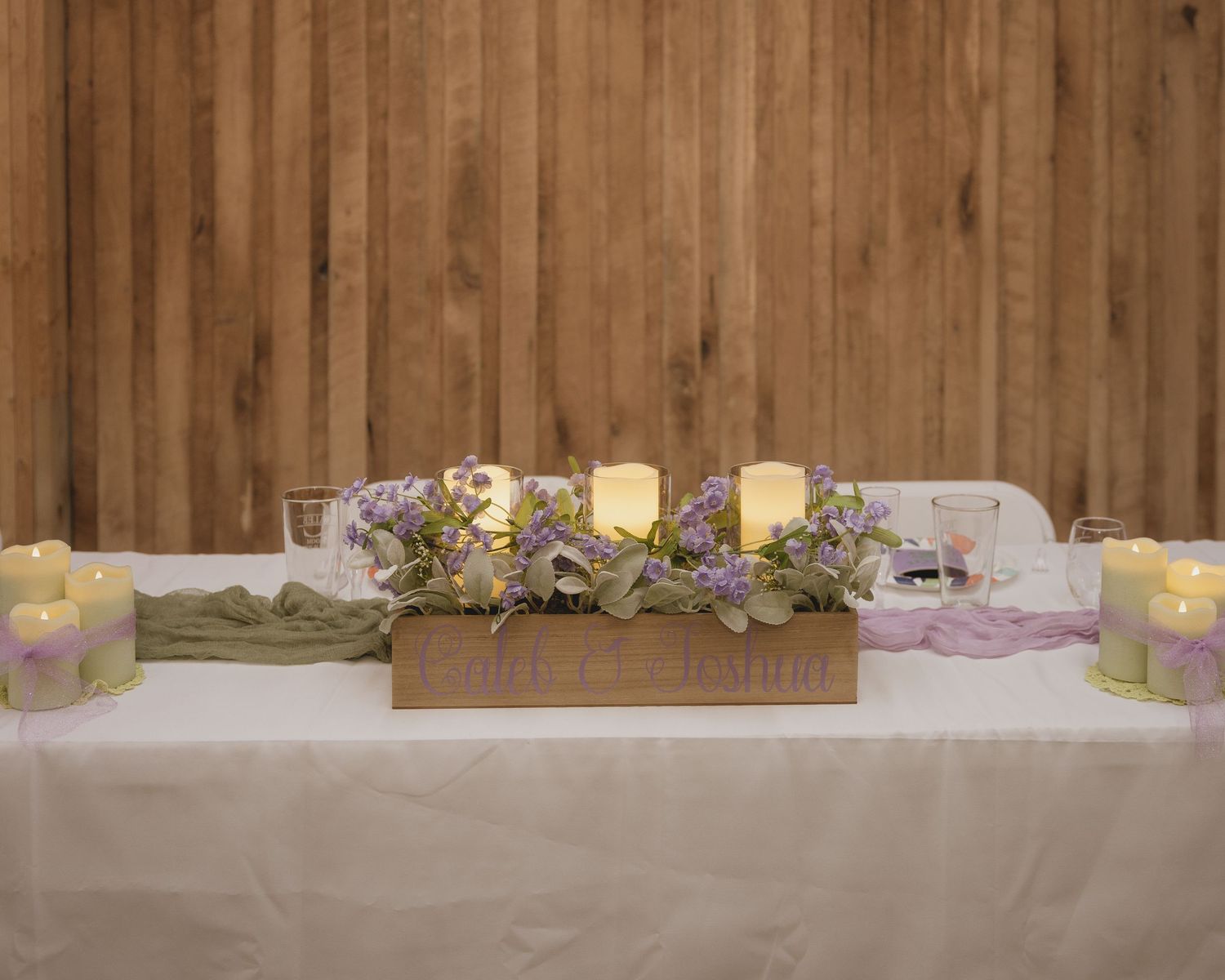 Elegant wedding head table with floral centerpiece, candles, and white tablecloth against a rustic wood wall.
