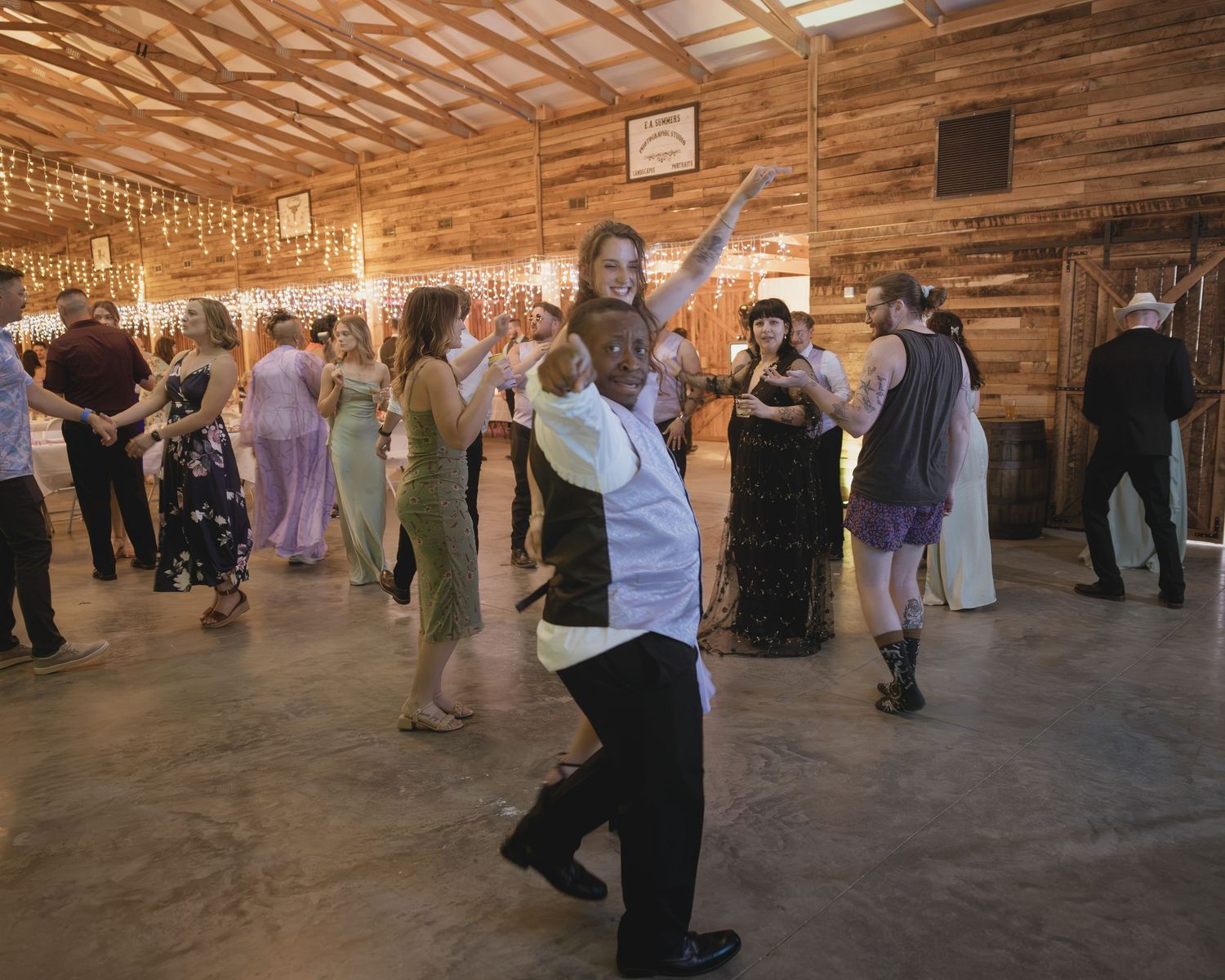 Diverse wedding guests dance joyfully on the floor of a warmly lit barn reception.