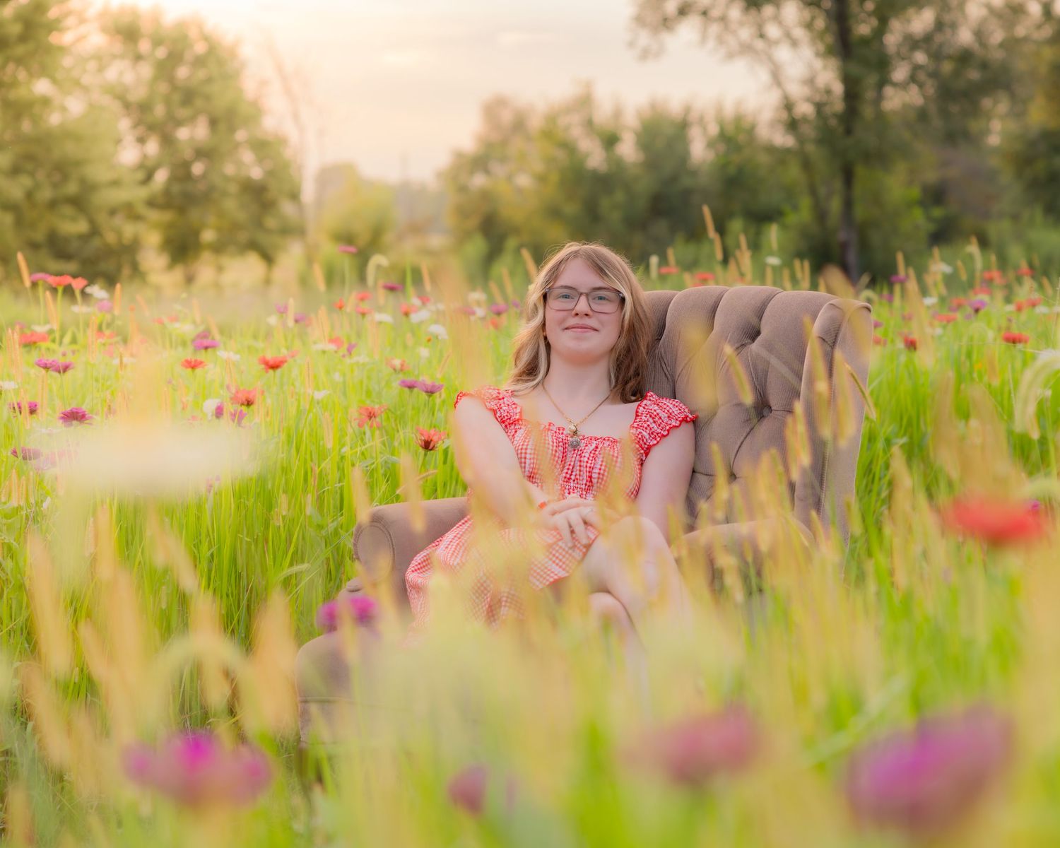 A young girl with glasses sits in a vintage chair surrounded by colorful wildflowers in a sunny meadow.