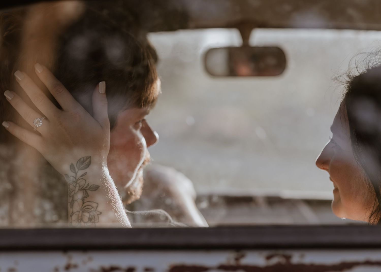 Couple sharing an intimate moment inside a vintage car, with tattoos and a ring visible through the window.