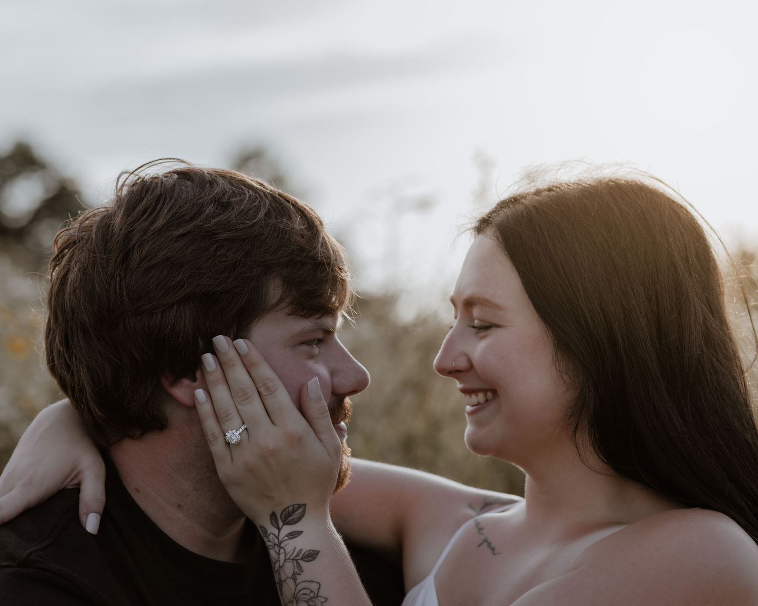 Couple smiling and laughing together outdoors at sunset, woman showing engagement ring on mans cheek.