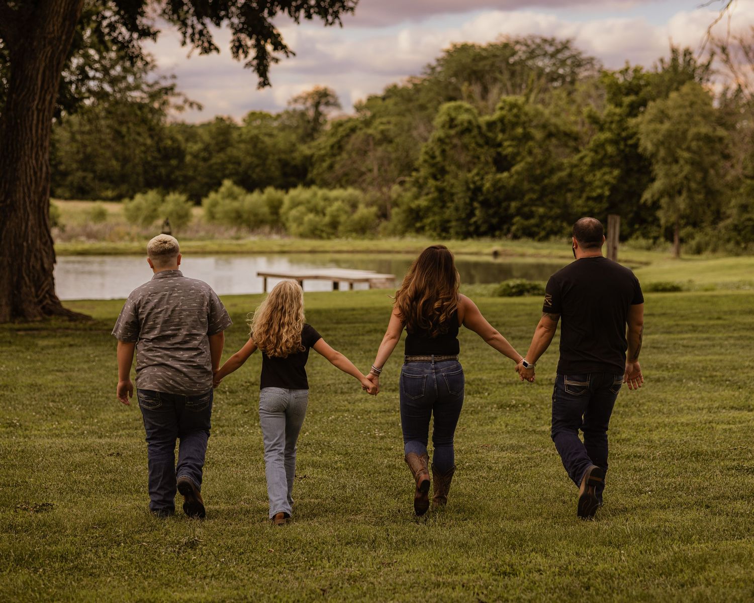 Family of four holding hands walking toward a serene pond in a lush green park setting.