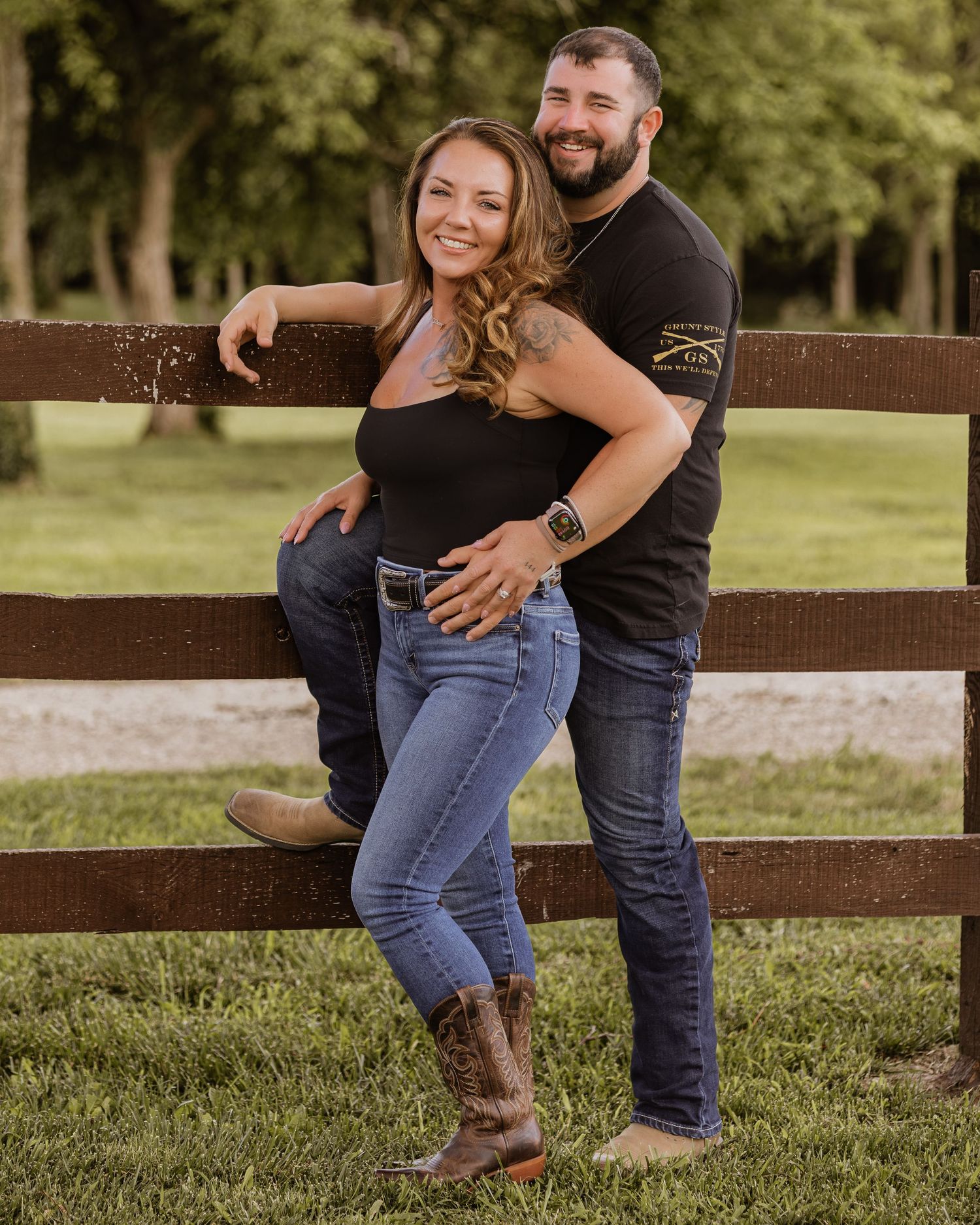 Smiling couple posing together against a rustic wooden fence on a sunny day at a farm.