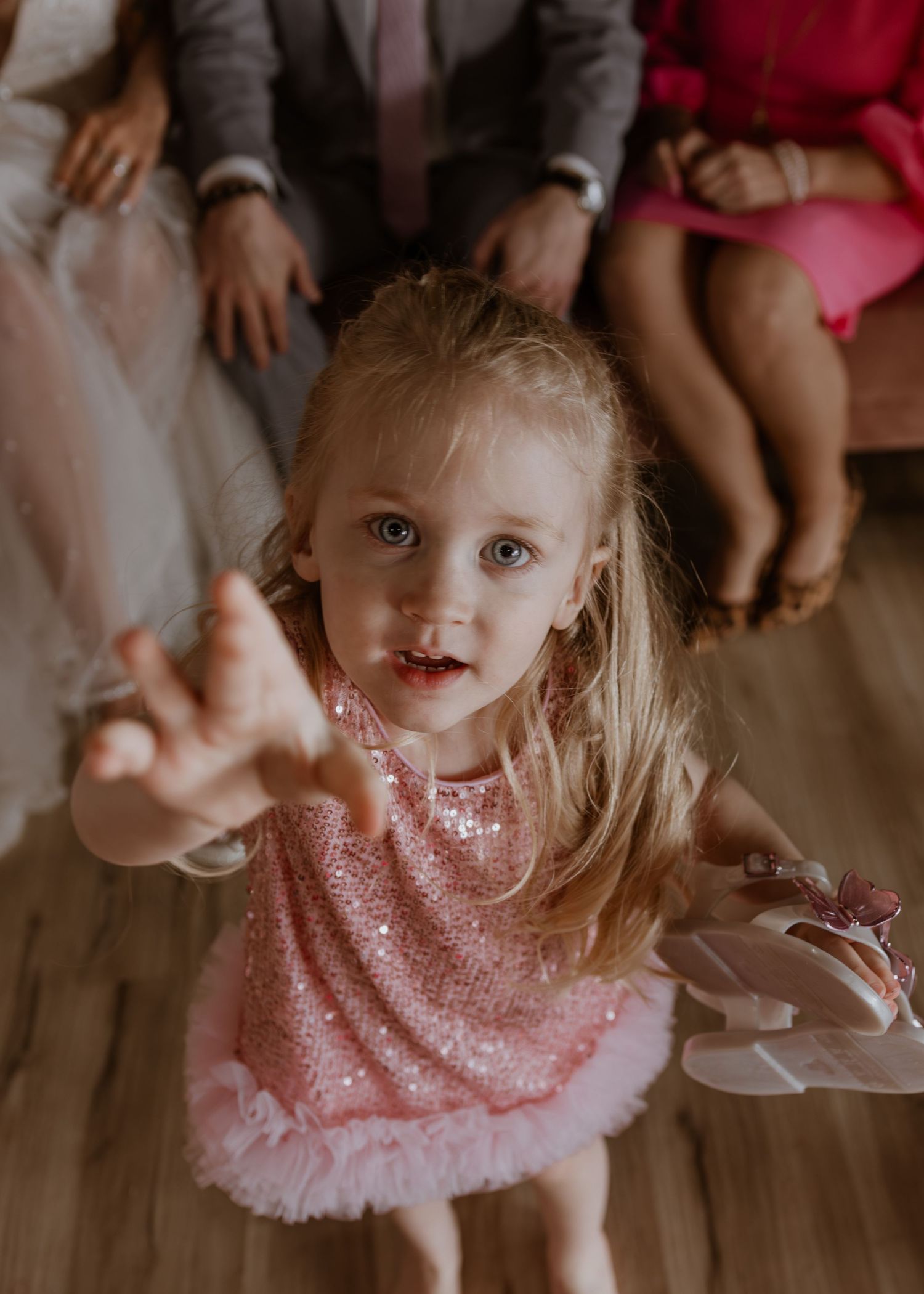 A toddler girl in a pink sequin dress reaches toward the camera at a wedding reception.