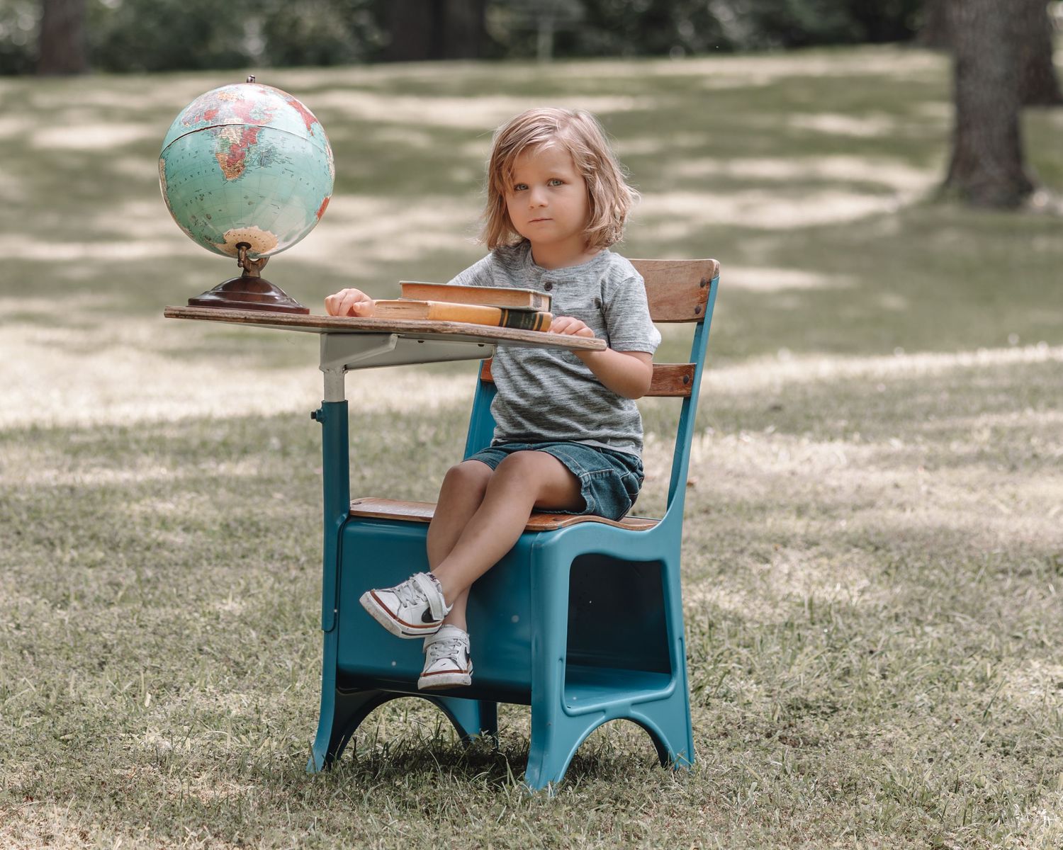 A child sits on a turquoise vintage school desk outdoors on grass while reading a book on a sunny day.