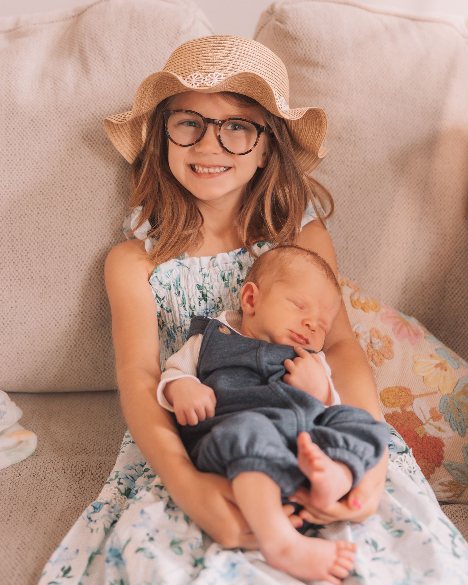 Young girl with glasses and straw hat smiling while holding a sleeping newborn baby in blue overalls.