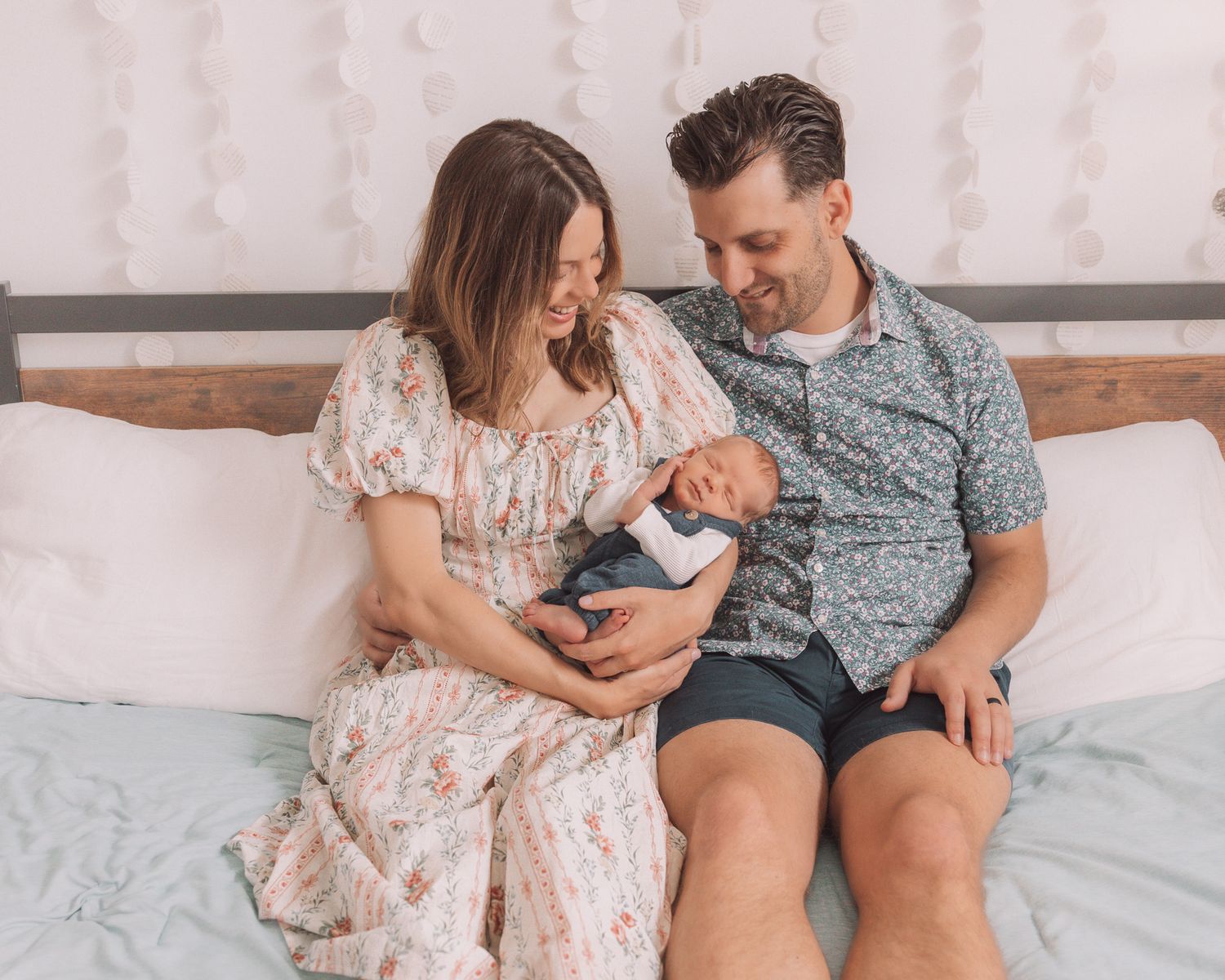 Couple sitting on a white bed smiling and cradling their newborn baby in a cozy bedroom setting.