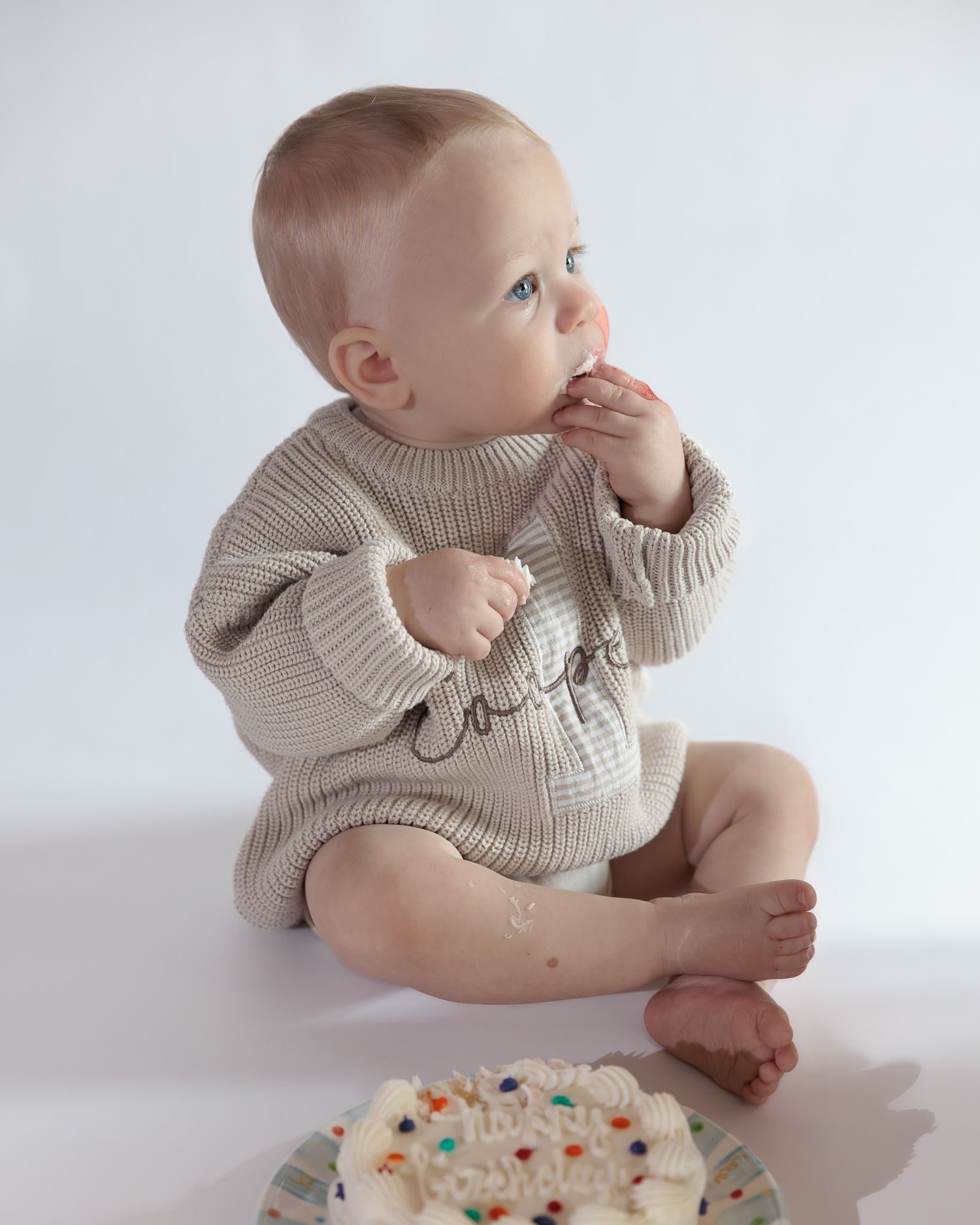 Baby boy in beige knit romper enjoys his first birthday cake smash, eating frosting with cake crumbles on white background.