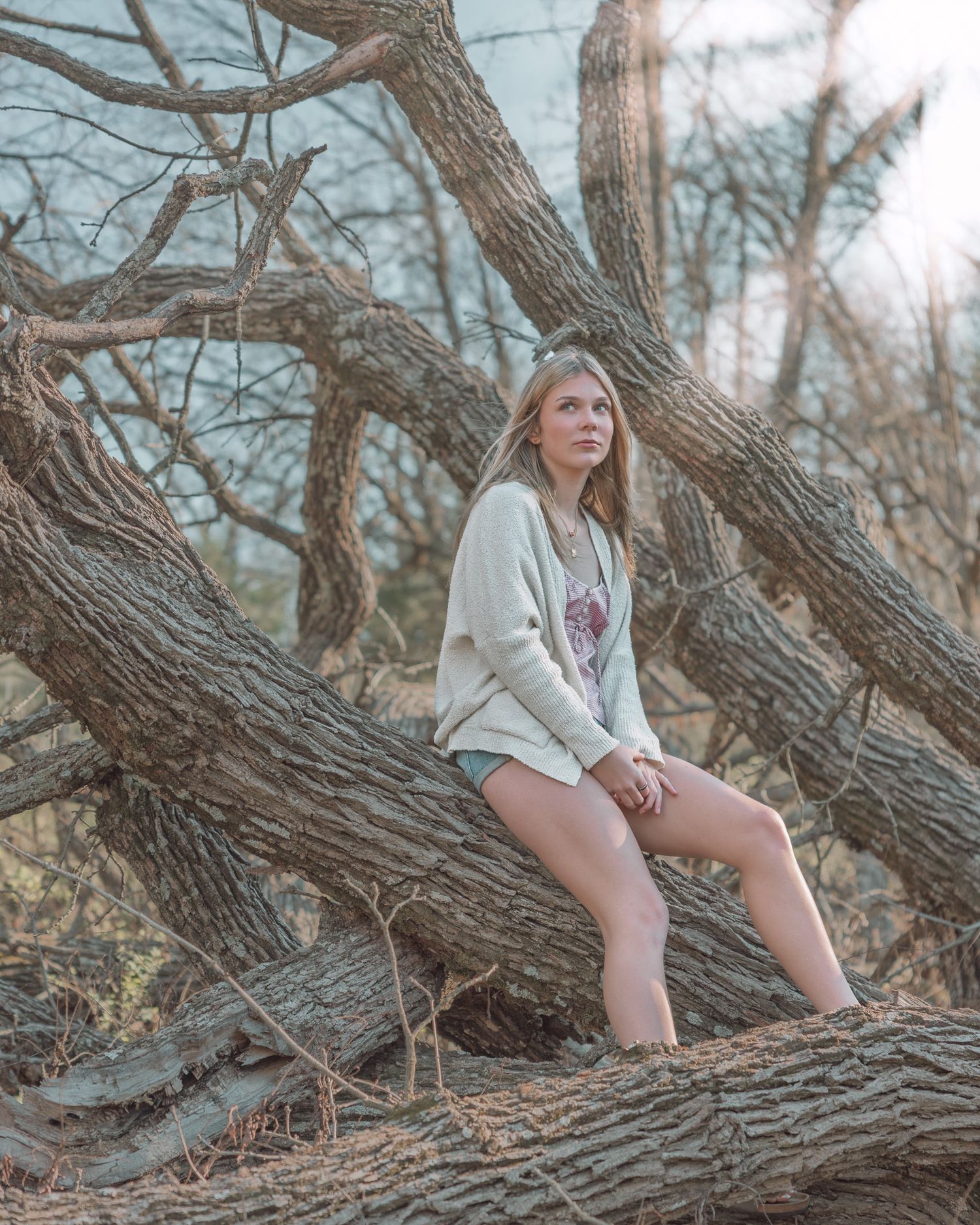 Young woman in white cardigan and denim shorts posing on fallen tree branches in a wooded outdoor setting.
