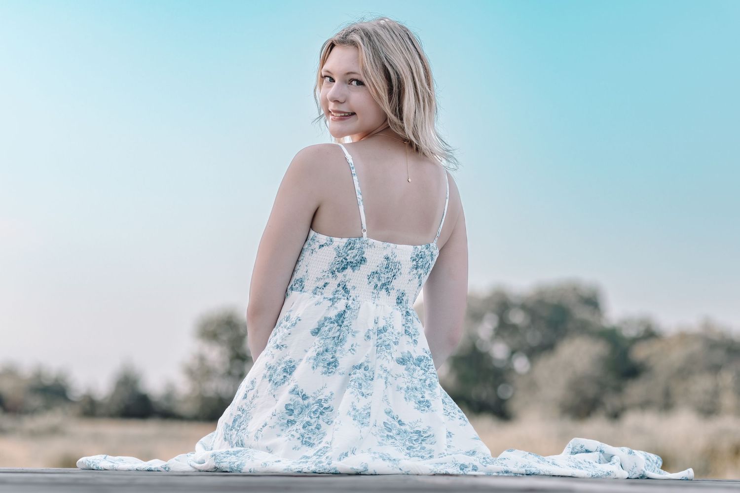 Person in white floral print dress poses gracefully in a sun-drenched outdoor setting with a natural background.
