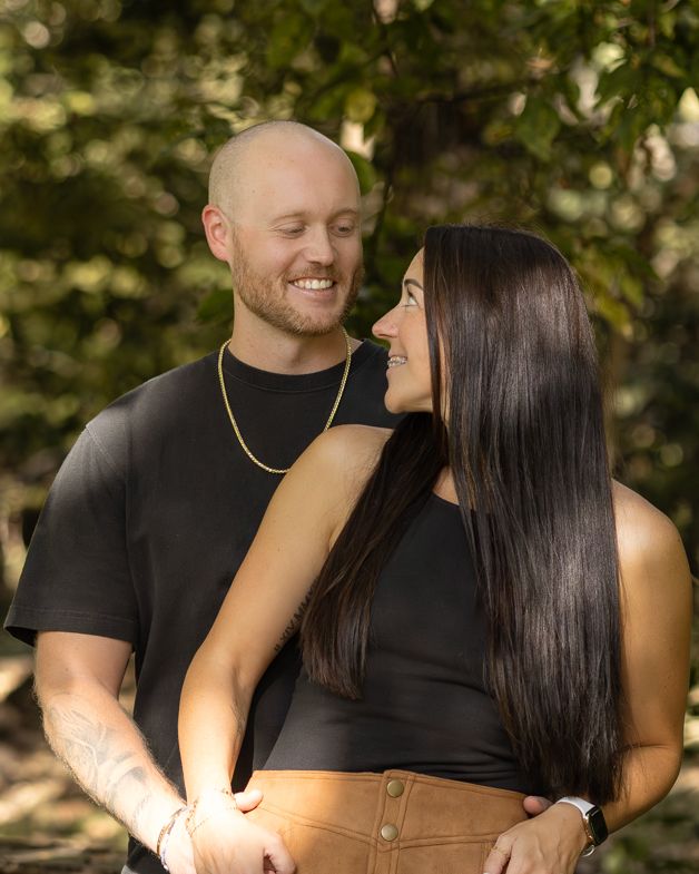 A smiling couple embraces outdoors among green trees, man in black tee with gold chain, woman with long dark hair in black crop top.