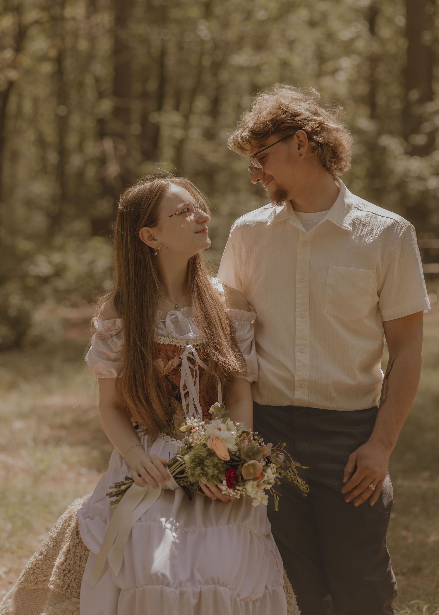 A couple in romantic attire shares a tender glance in a sunlit forest, woman holding a bouquet.