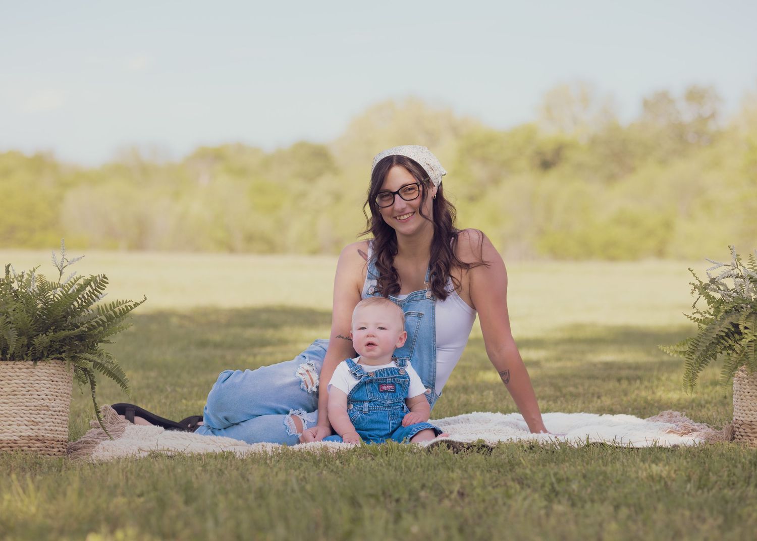 A mother and baby sit on a blanket in a sunny green field, both smiling in blue outfits.
