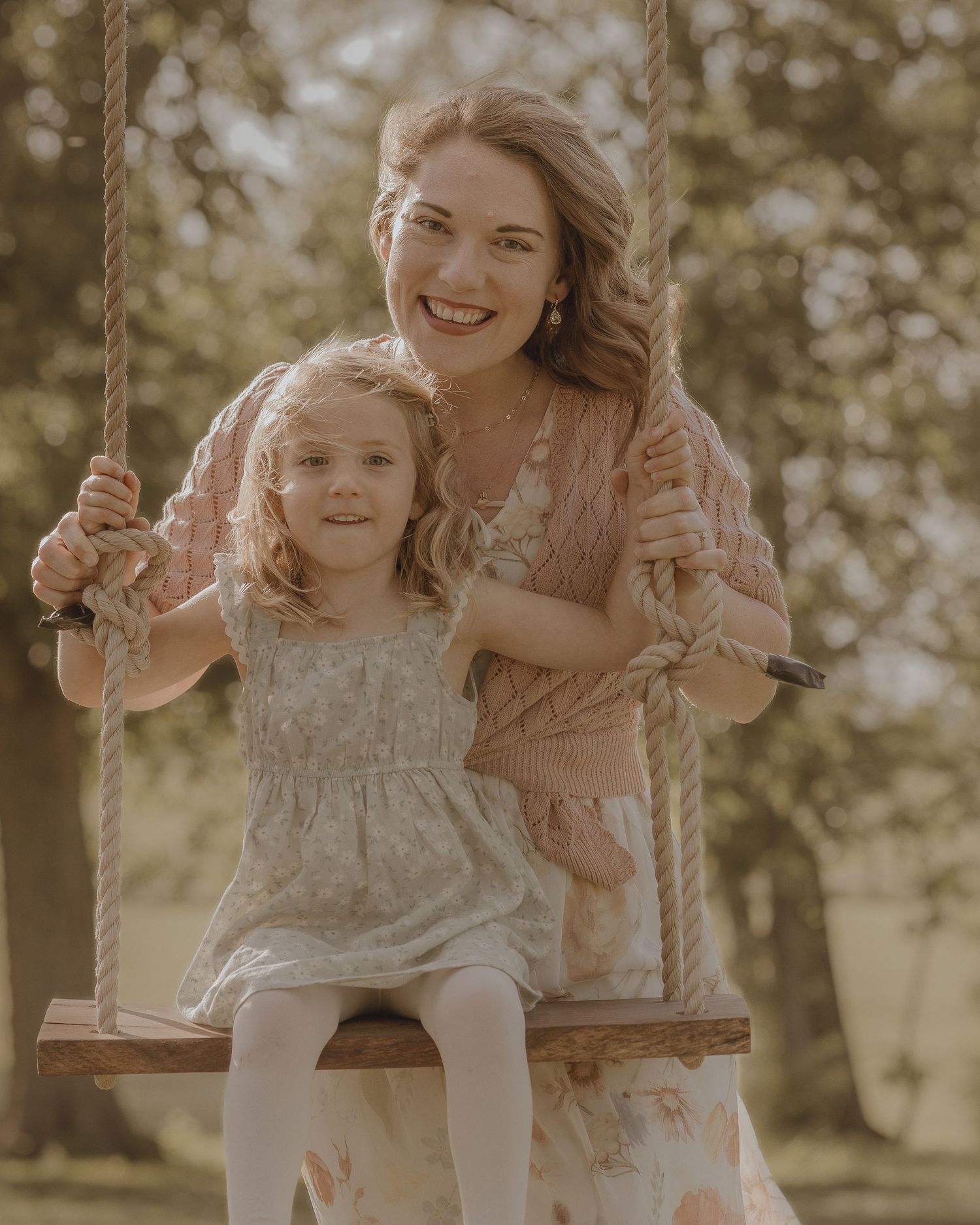 A smiling woman and young girl in floral dresses pose together on a wooden rope swing outdoors.