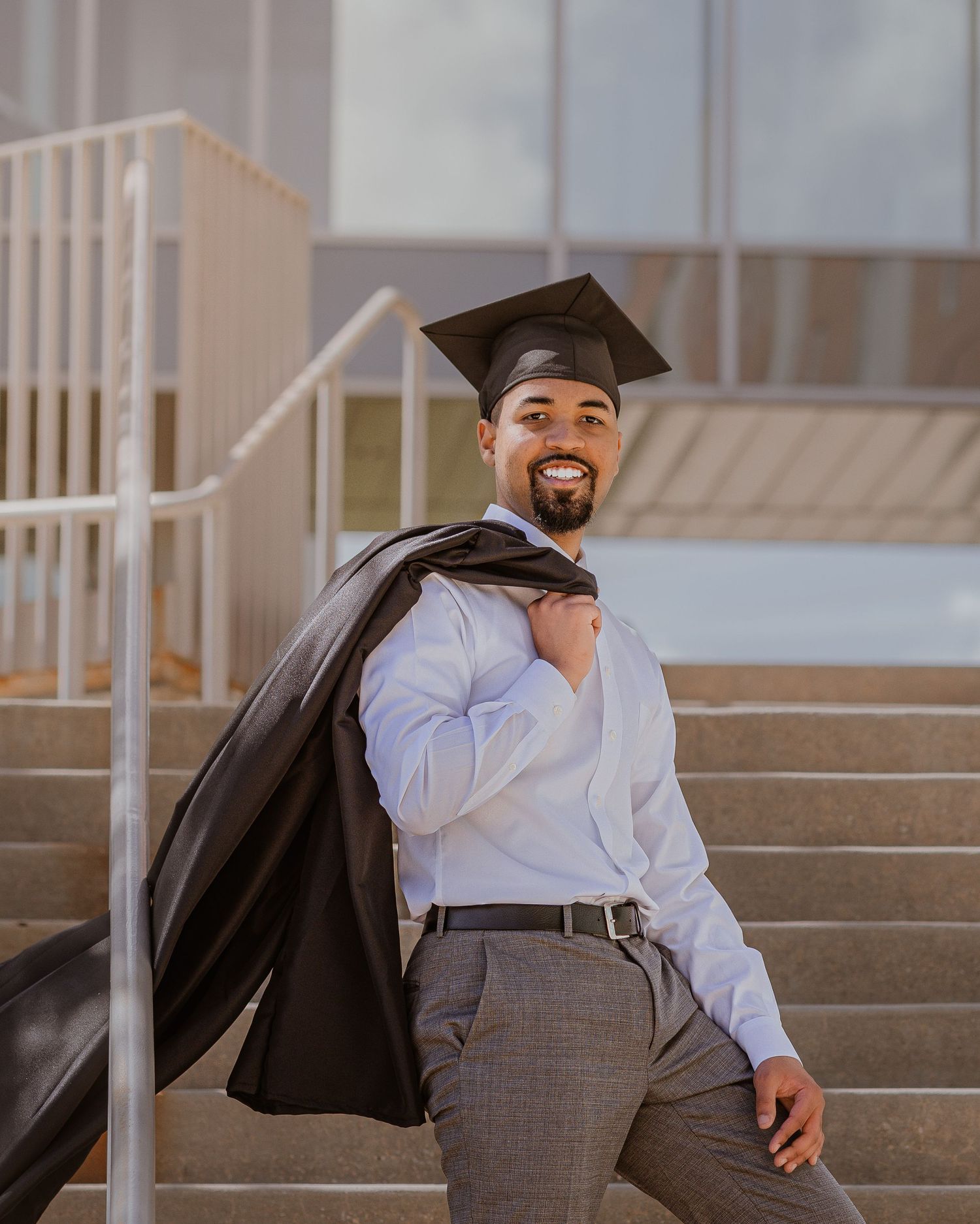 Smiling graduate in cap and gown draping over shoulder poses on outdoor steps in white shirt and gray pants.