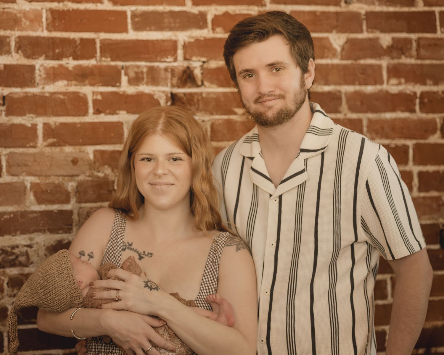 Young couple smiling and holding a newborn baby in front of a rustic brick wall backdrop.