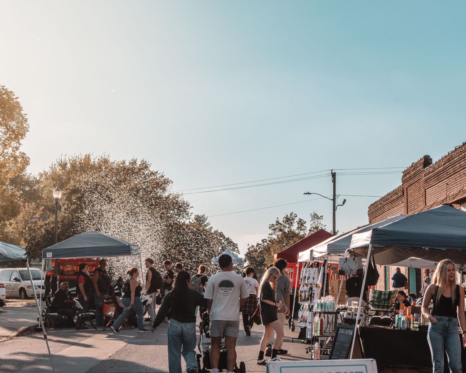 People browse vendor booths at an outdoor street market with pop-up tents on a sunny autumn day.