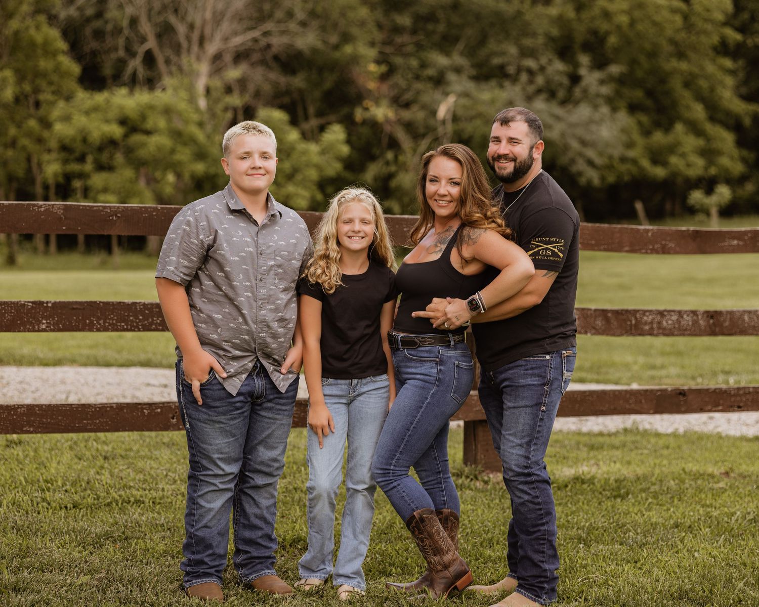 Family of four smiling outdoors by a wooden fence on a green lawn with trees in the background.