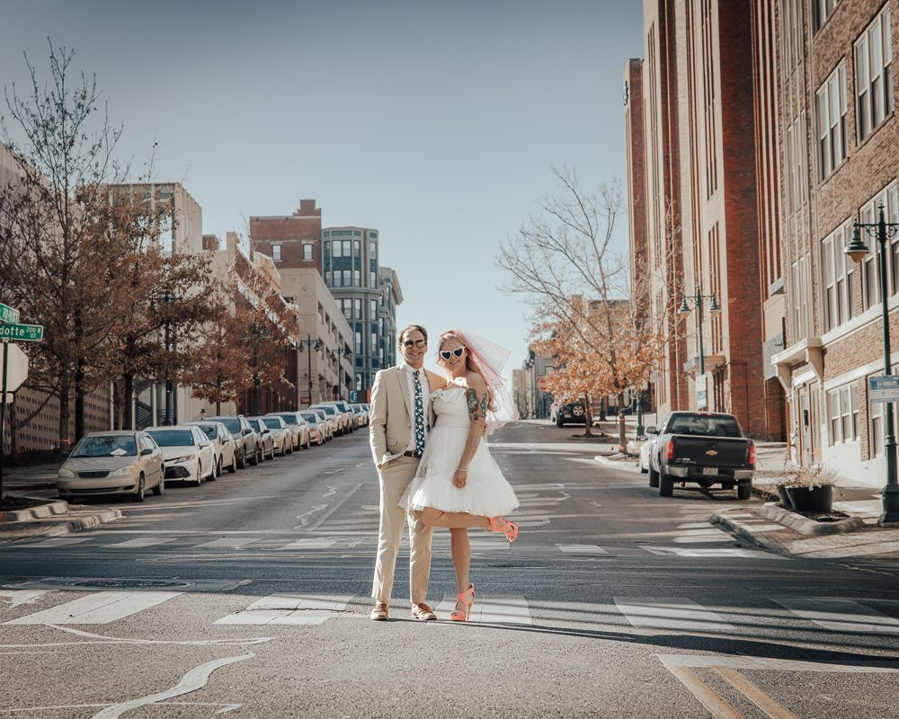 A couple in light-colored outfits stands together in the middle of an urban street with tall buildings.