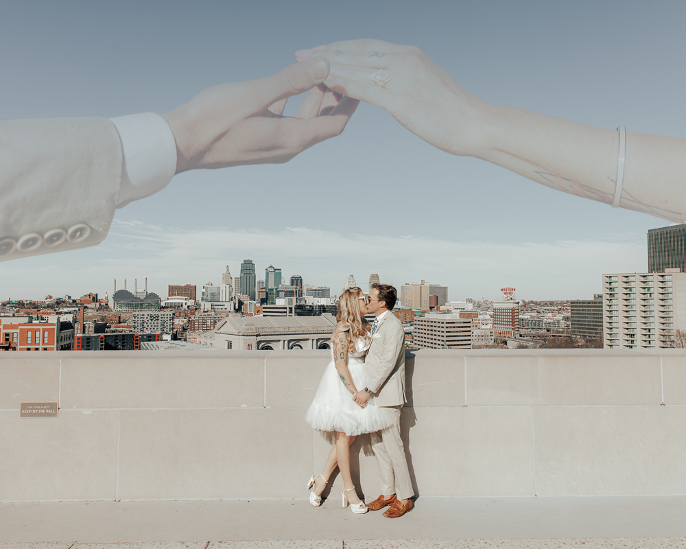 A couple embraces on a rooftop with hands forming an arch above them against a cityscape backdrop.